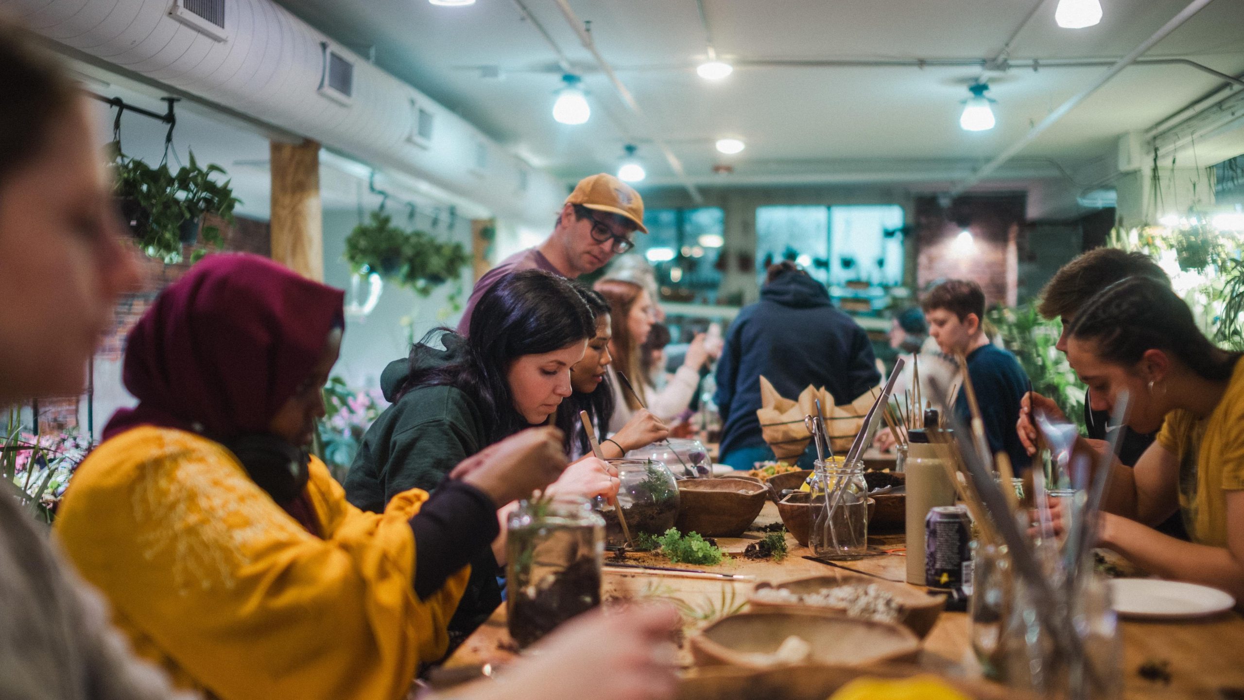 A group of USM students gather around a long wooden table building terrariums together inside a warmly lit plant shop, with hanging greenery and supplies scattered across the table.