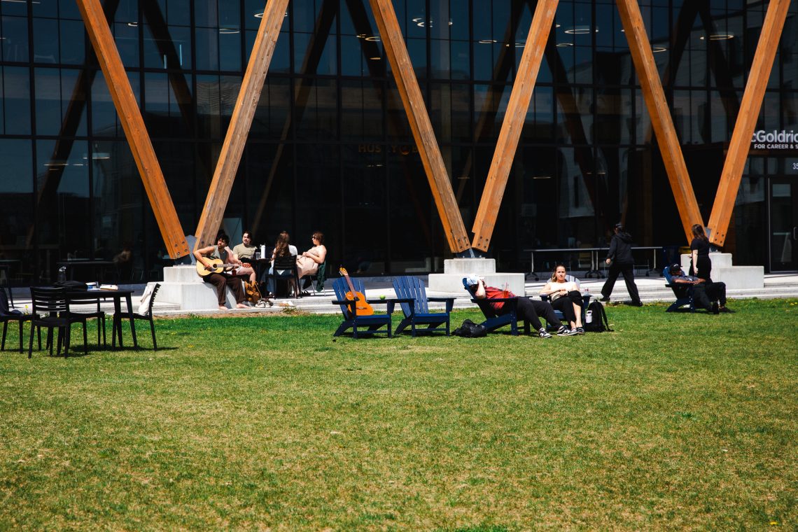Students gathered on the Bean Green outside McGoldrick Center, some sitting in Adirondack chairs while others play guitar and relax on the lawn