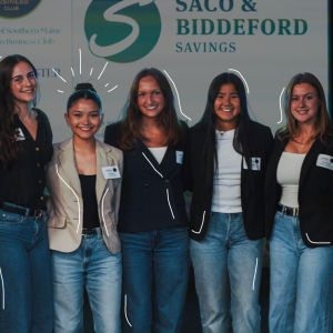 Seven students stand together in front of a University of Southern Maine Women in Business Club banner and a Saco and Biddeford Savings sign at a networking event.