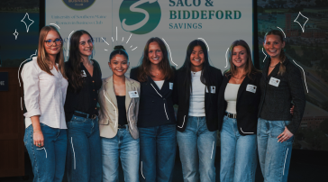 Seven students stand together in front of a University of Southern Maine Women in Business Club banner and a Saco and Biddeford Savings sign at a networking event.