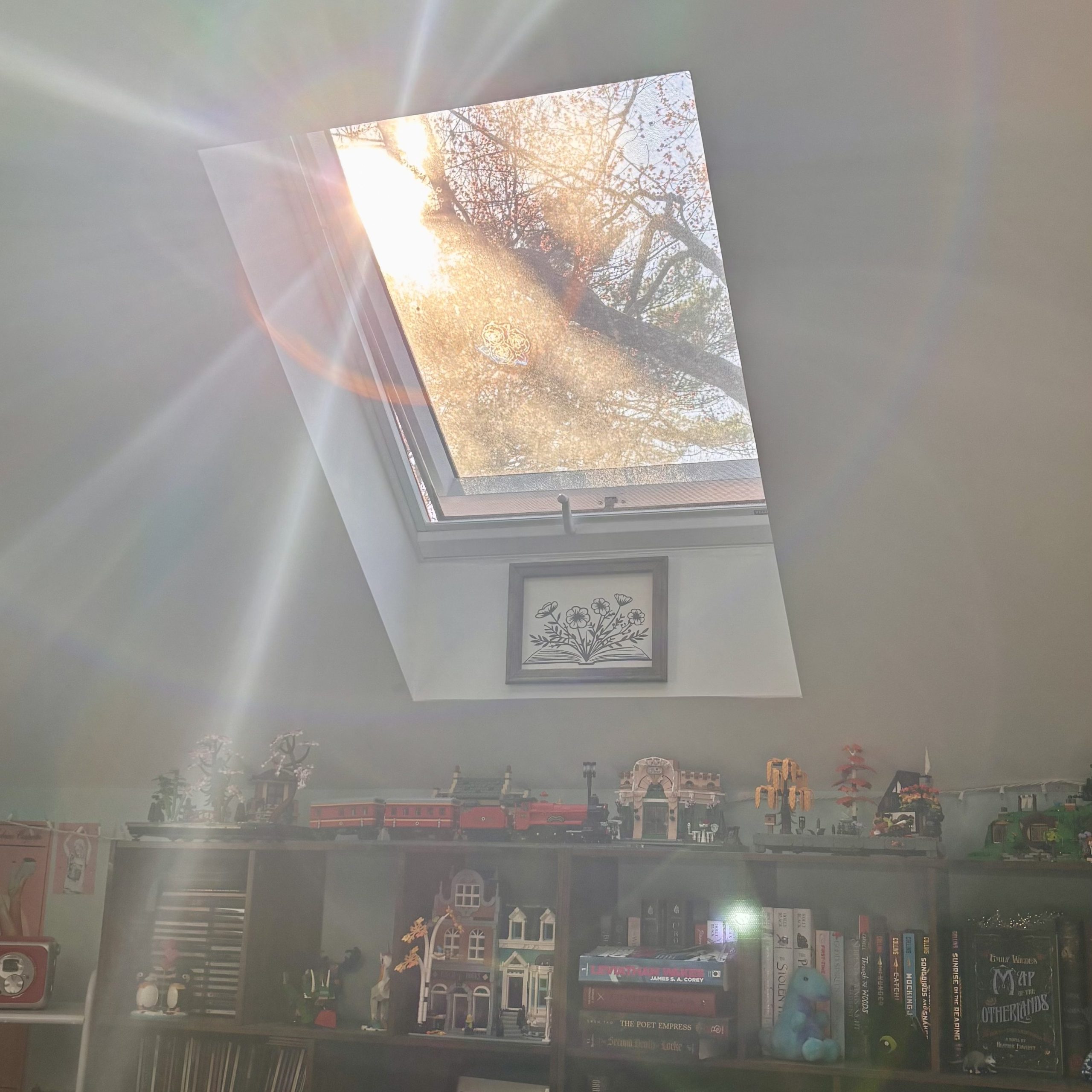 Sunlight streams through a skylight, casting rainbow light across two bookshelves filled with books, figurines, and LEGO sets in a cozy bedroom.