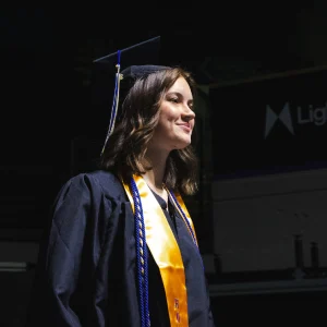 A graduate in a cap, gown, and gold USM stole stands in the staging area at Cross Insurance Arena, looking ahead before the processional begins.