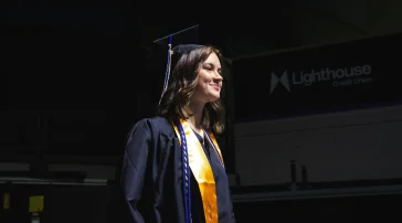 A graduate in a cap, gown, and gold USM stole stands in the staging area at Cross Insurance Arena, looking ahead before the processional begins.