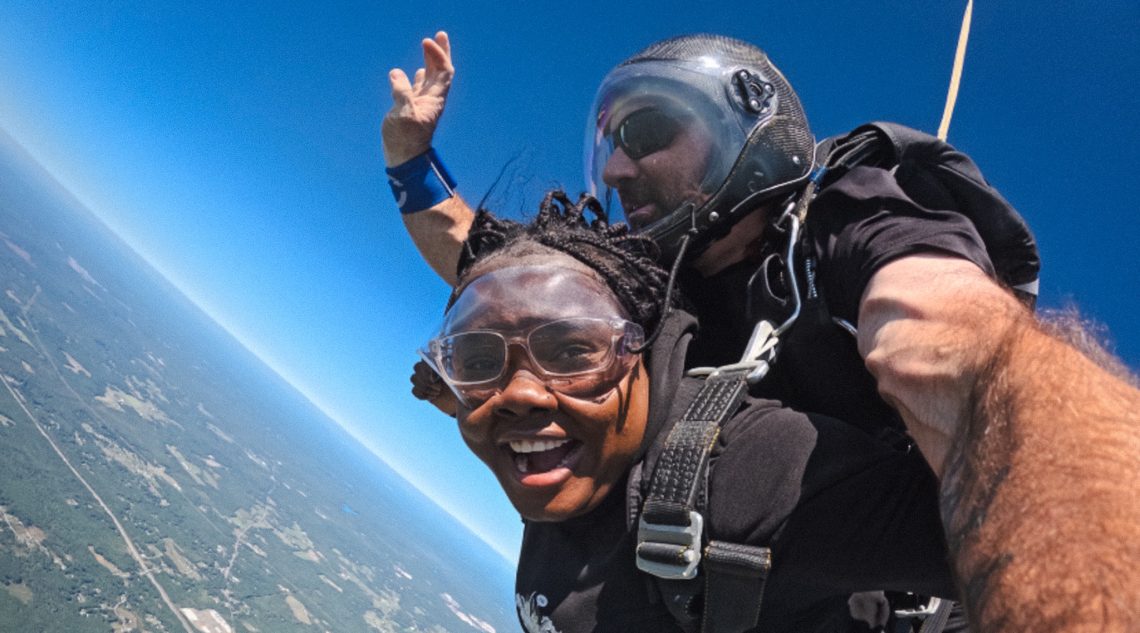 A USM student smiles widely mid-tandem skydive with her instructor, with bright blue sky above and the curve of the Maine landscape far below.