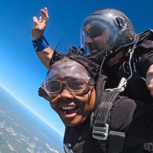 A USM student smiles widely mid-tandem skydive with her instructor, with bright blue sky above and the curve of the Maine landscape far below.
