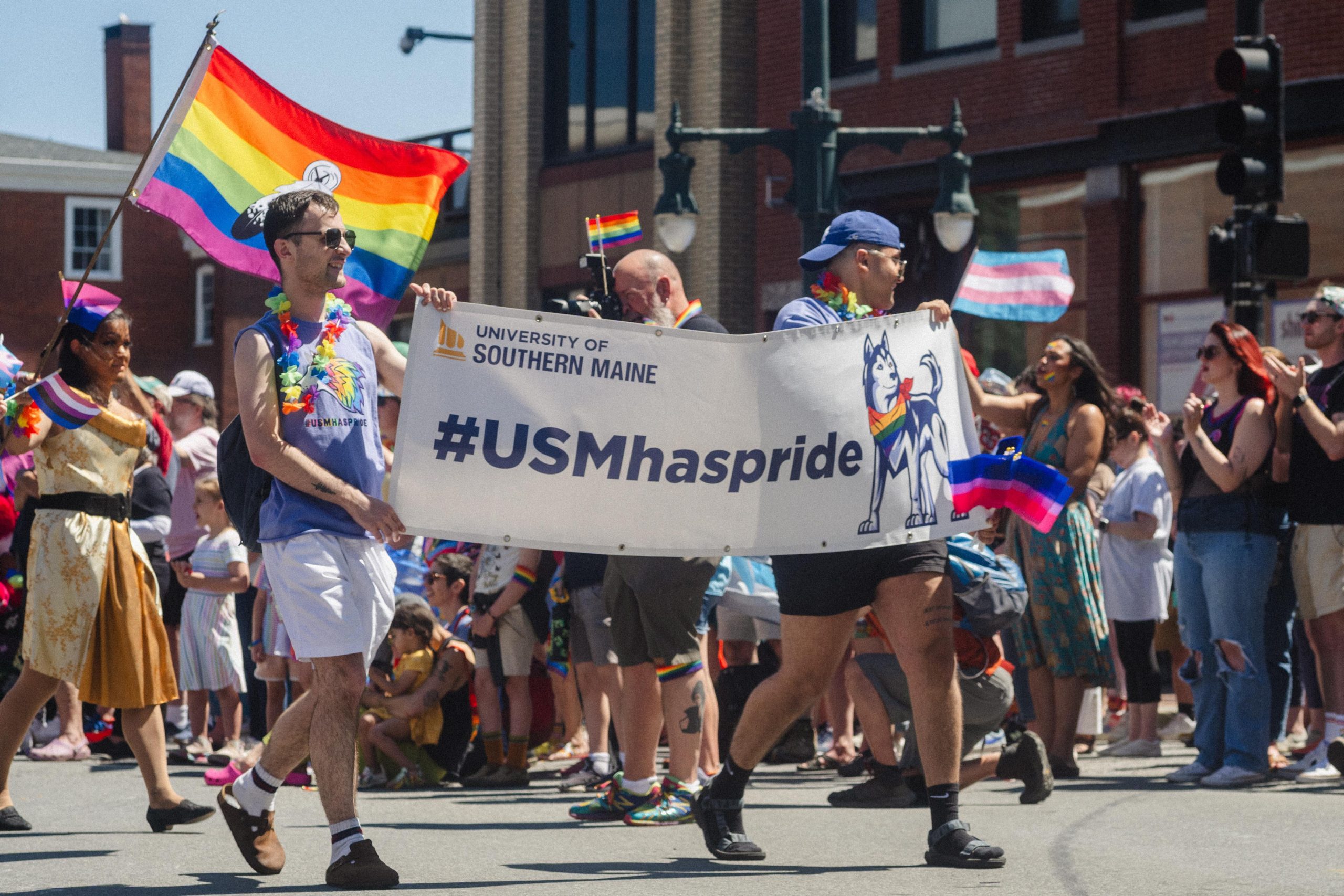 USM community members marching in Pride Portland parade carrying a banner reading "University of Southern Maine #USMhaspride" with rainbow flags and lei