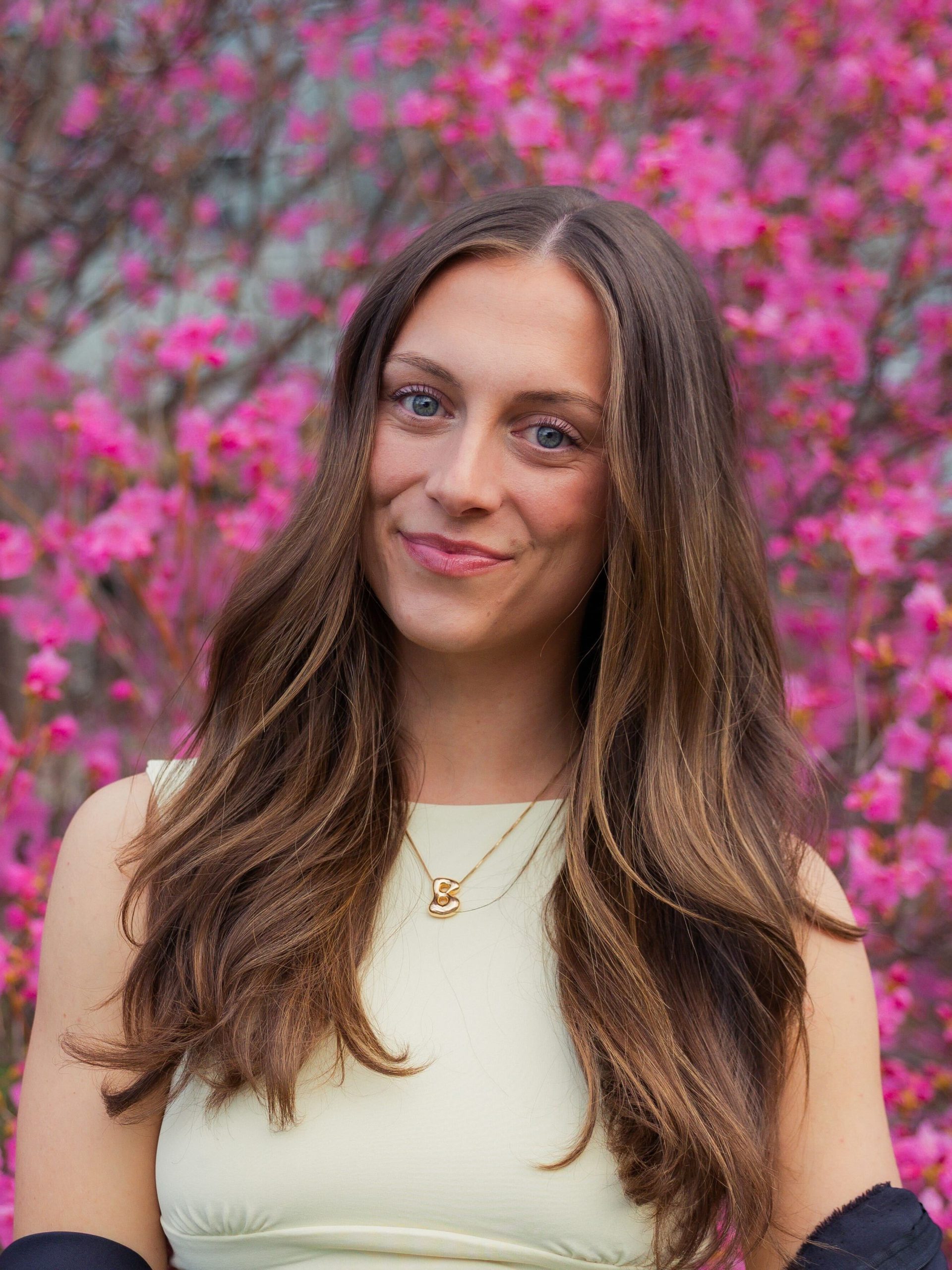 Bridgette Toland smiles in front of a flowering pink tree in full bloom, wearing a pale yellow top and a gold initial necklace.