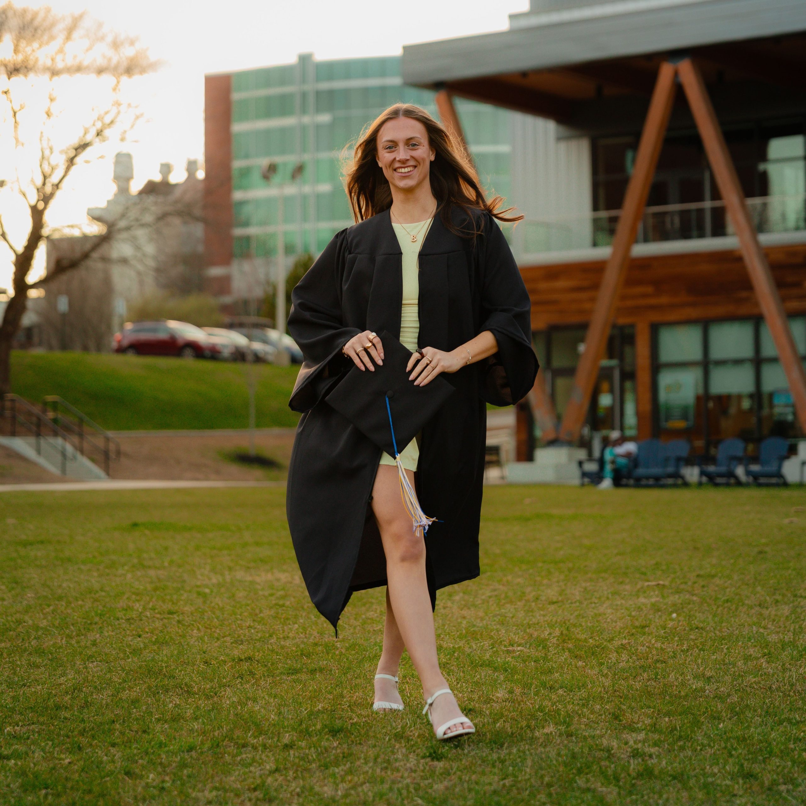 Bridgette Toland walks across the green lawn of USM's Portland campus in her graduation gown, holding her cap and smiling, with the McGoldrick Center for Career and Student Success visible in the background.