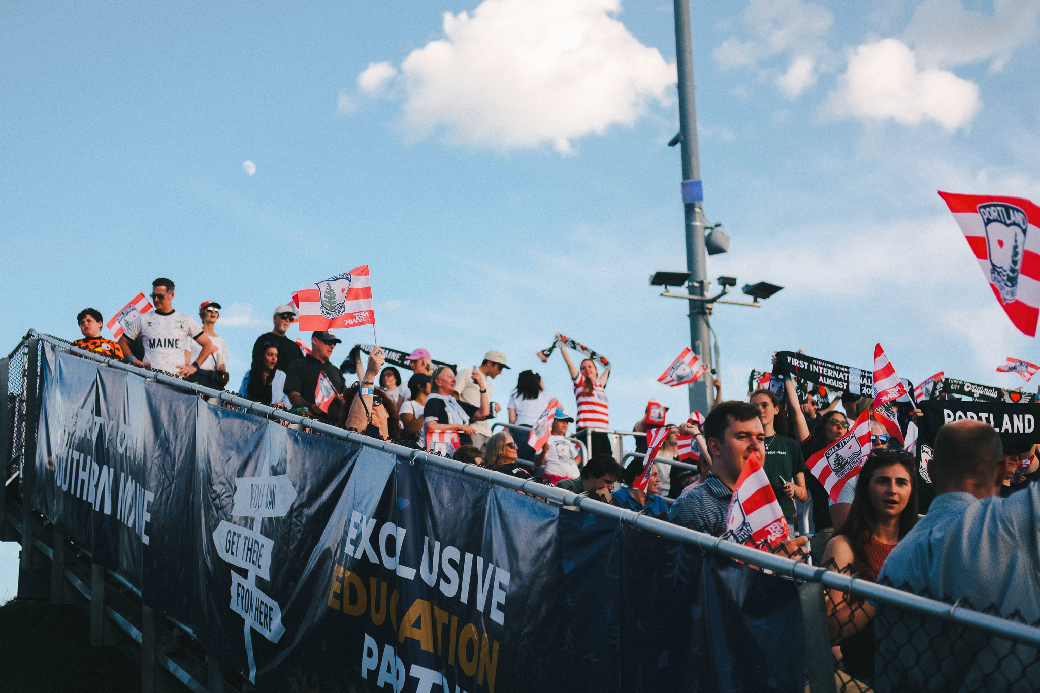 Hearts of Pine soccer fans waving red and white flags next to the USM exclusive education partner banner at Fitzpatrick Stadium in Portland, Maine