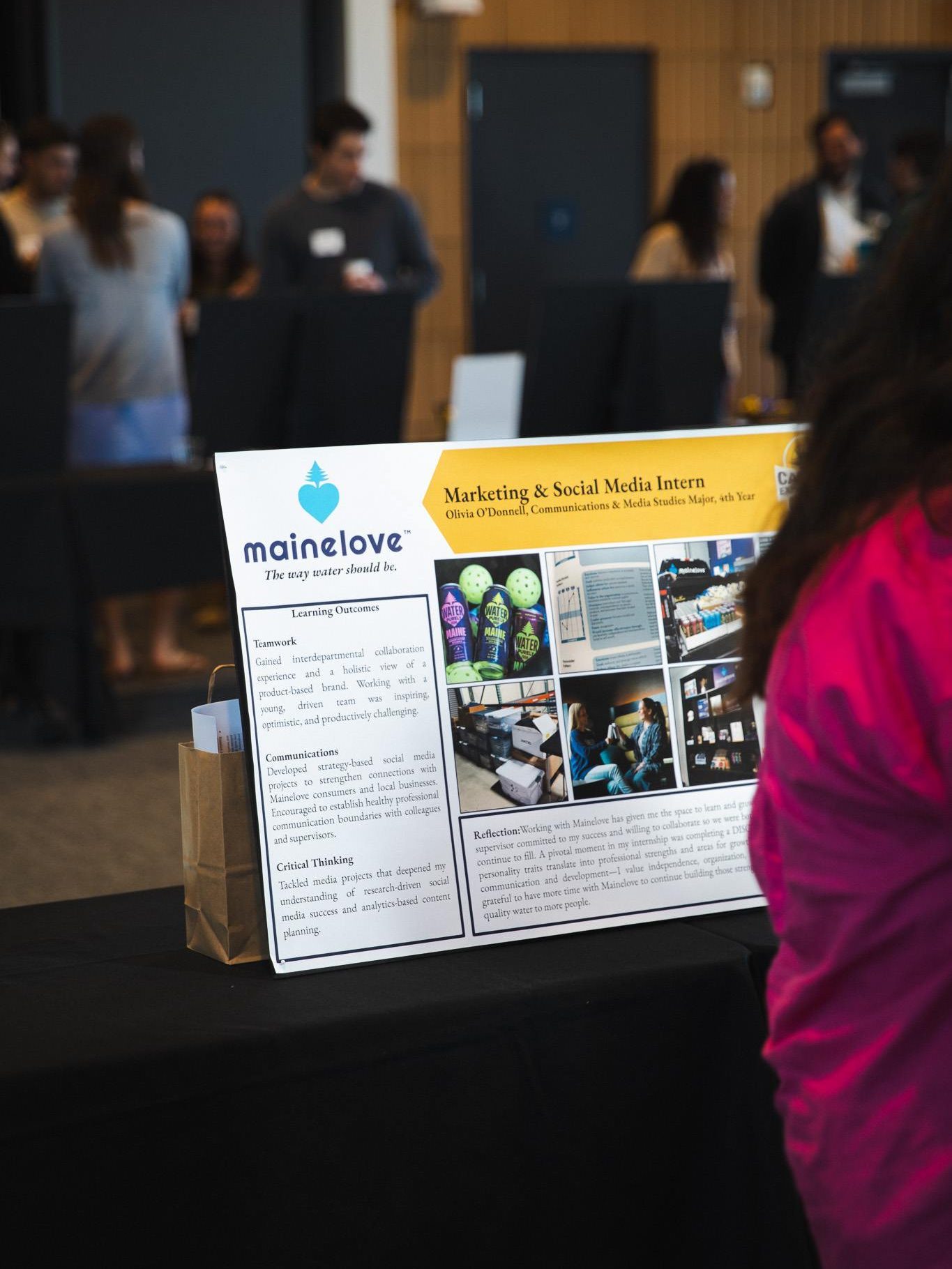 Olivia O'Donnell's CEIP poster for her mainelove internship on display at the CEIP Spring Poster Exhibition, with attendees visible in the background.