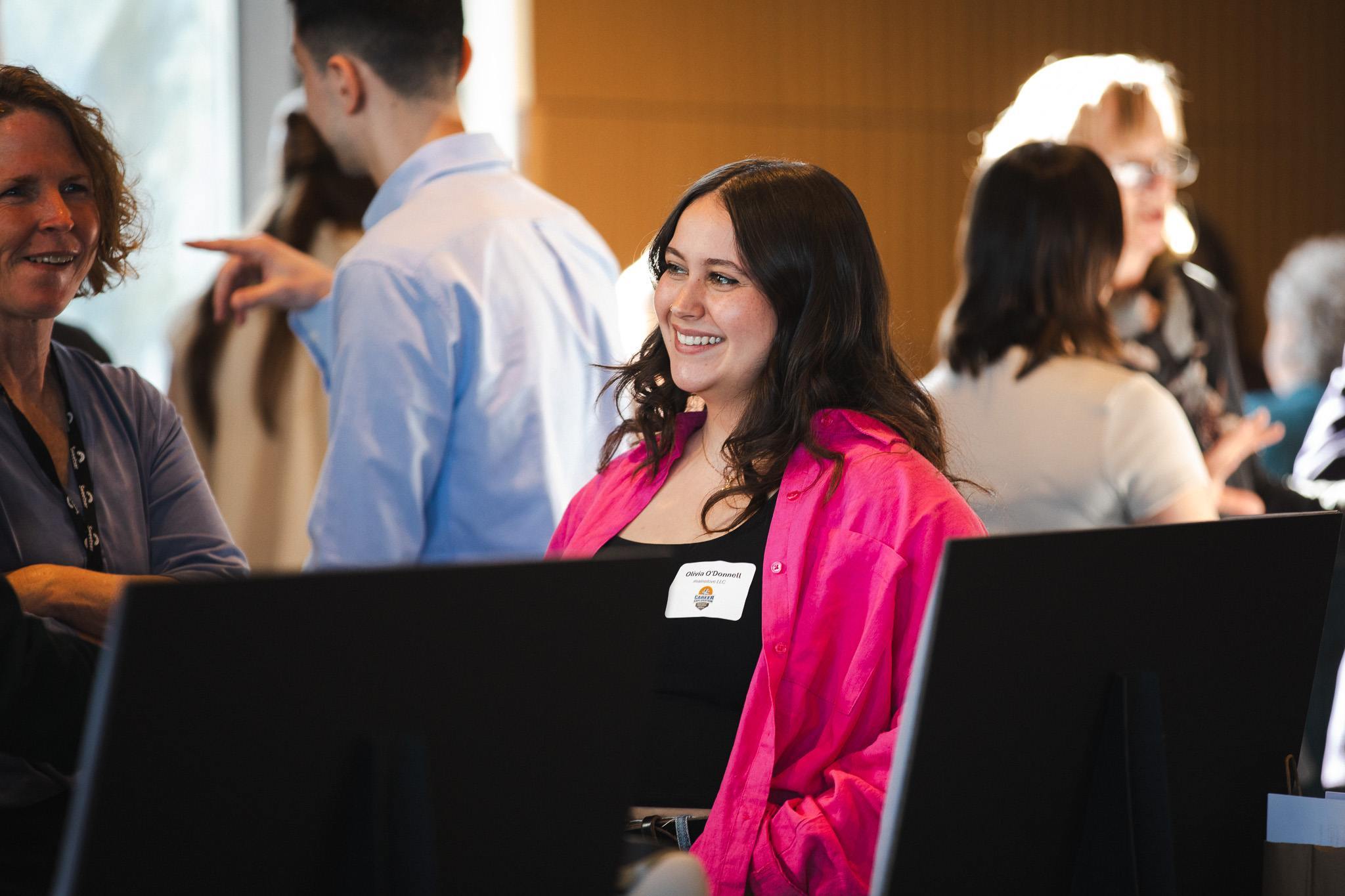 Olivia O'Donnell smiles during the CEIP Spring Poster Exhibition, wearing a bright pink blazer and a name tag, surrounded by other attendees.