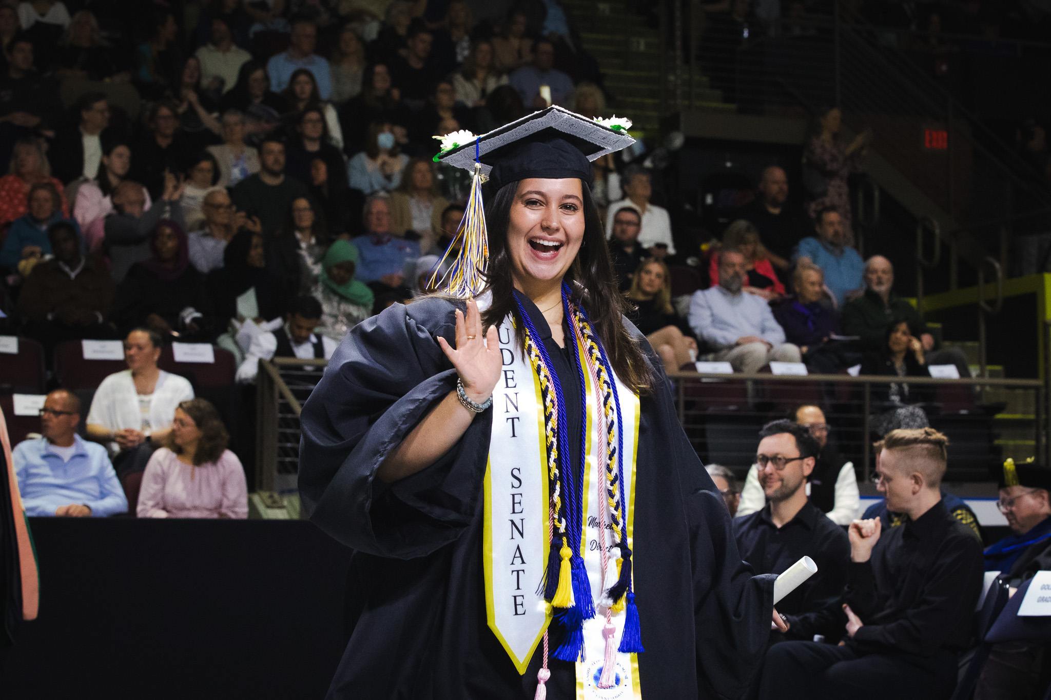 Olivia O'Donnell waves and smiles during USM's commencement ceremony at Cross Insurance Arena, wearing her cap, gown, and Student Senate stole.