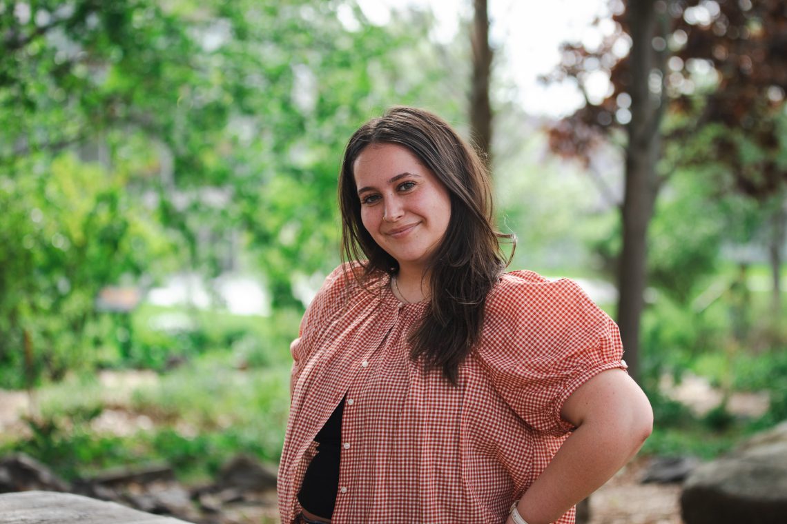 Olivia O'Donnell smiles outdoors on the USM campus, wearing a red gingham top, with green trees in the background.