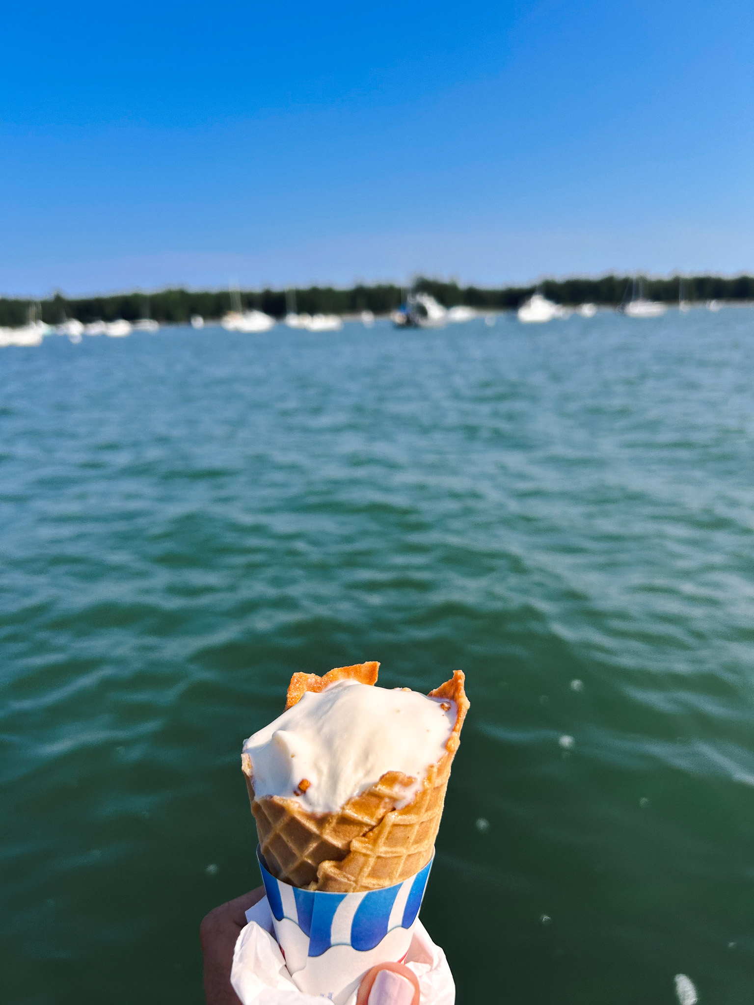 Hand holding an ice cream cone in a waffle cone with sailboats and harbor water in the background, Maine