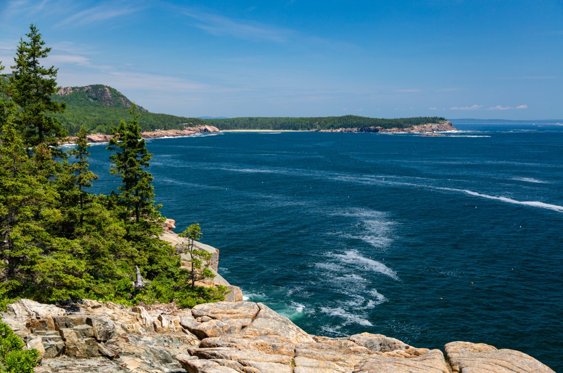 Rocky Maine coastline with evergreen trees, granite cliffs, and the Atlantic Ocean stretching to the horizon with islands visible in the distance