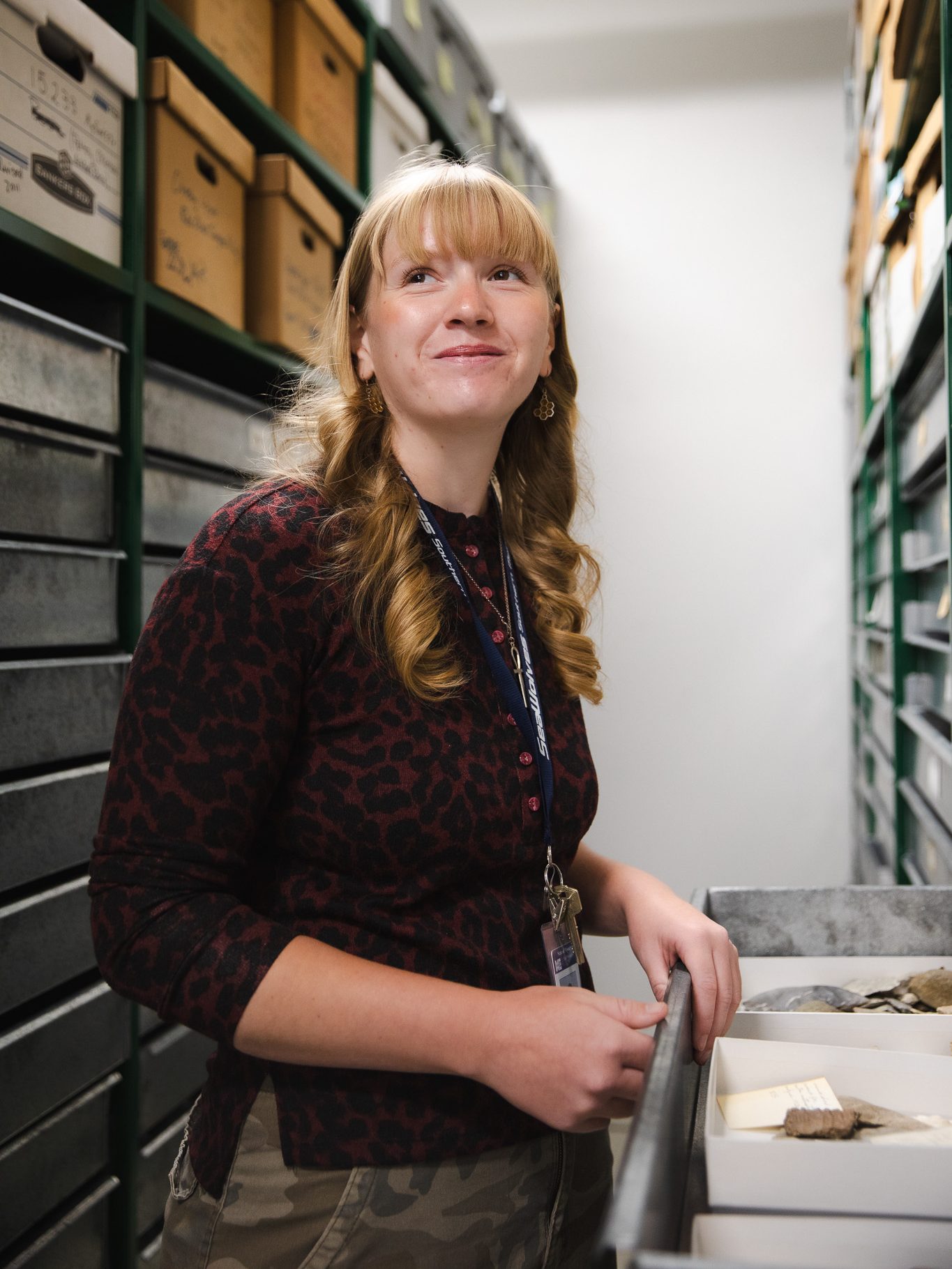 Camdyn Johnson stands among archival storage shelves in the Maine State Museum's collections building, opening a drawer of artifacts while smiling.
