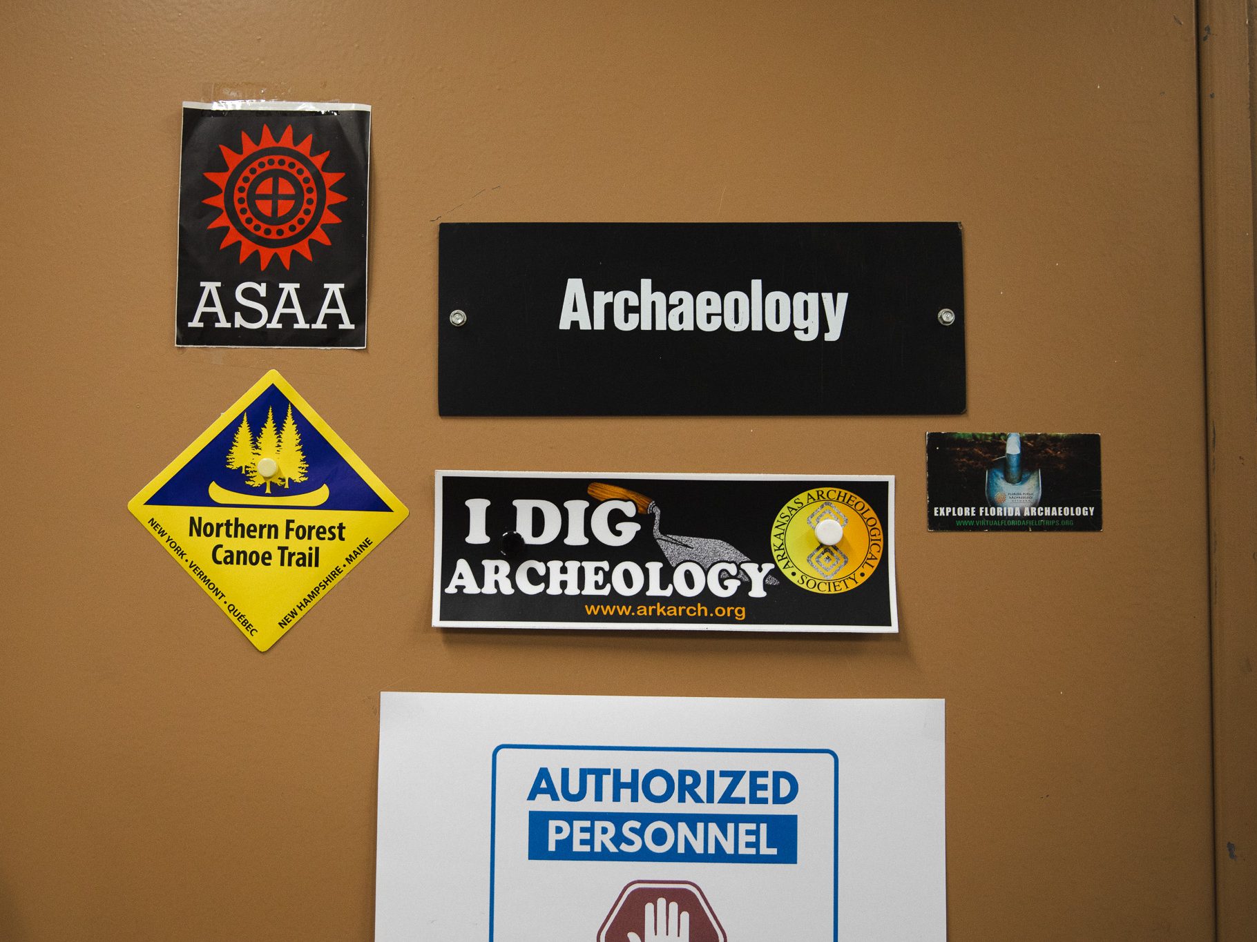 A door labeled "Archaeology" at the Maine State Museum, decorated with stickers reading "I Dig Archeology," "Northern Forest Canoe Trail," and an "Authorized Personnel" sign.