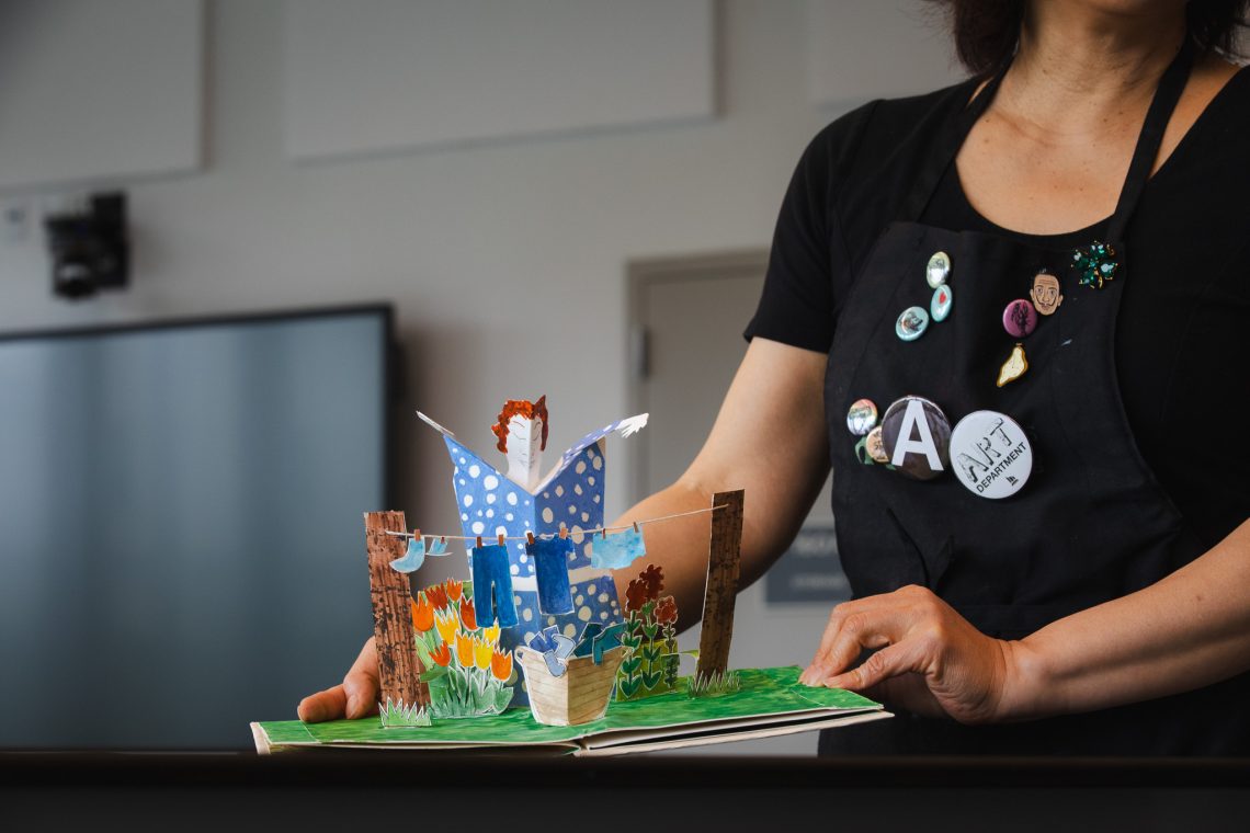 Annie Lee-Zimerle holds open a colorful pop-up artist book featuring a three-dimensional garden scene.