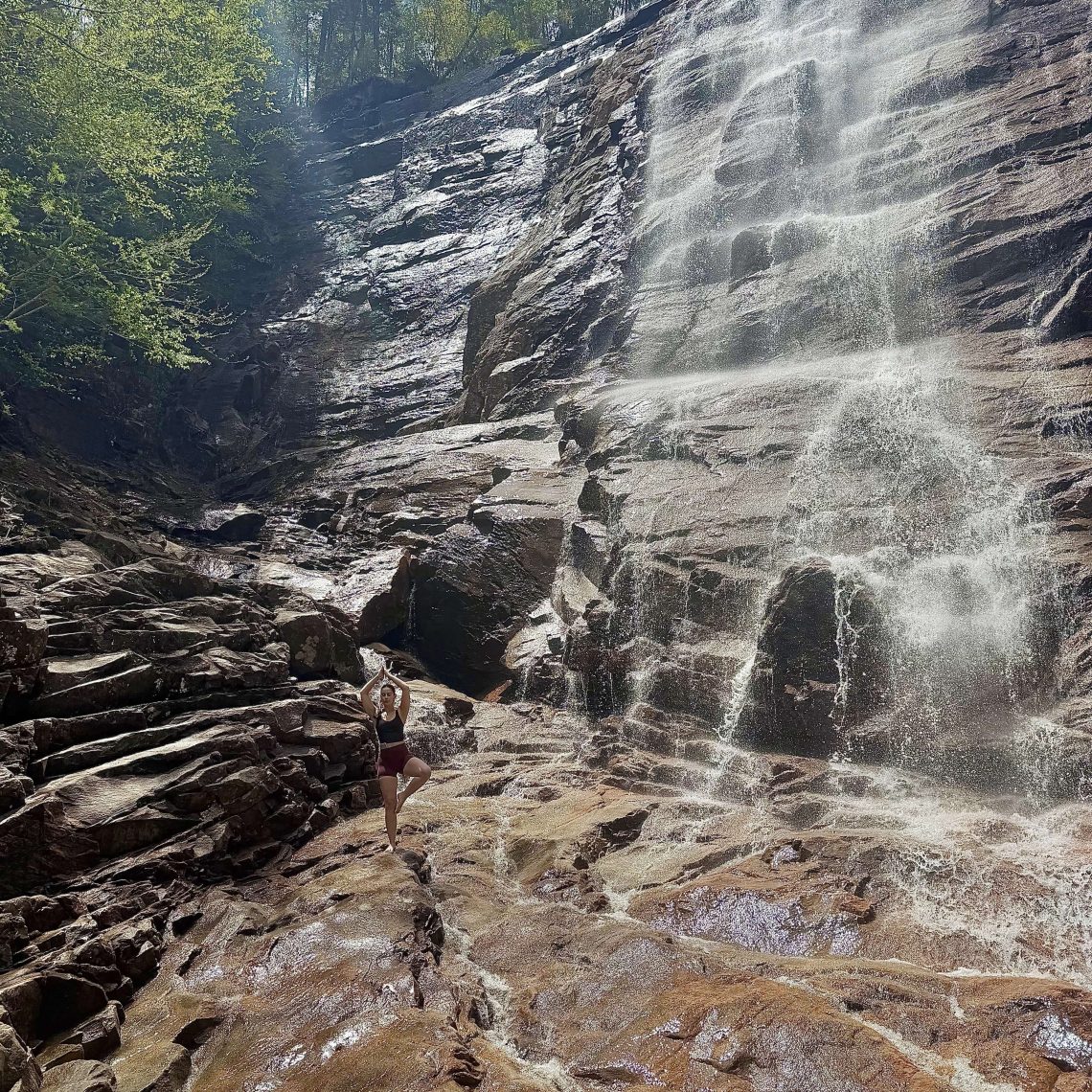 A student strikes a tree pose yoga position on the rocky base of Arethusa Falls in Crawford Notch, NH. The massive waterfall cascades down a wide granite rock face behind her, surrounded by green trees under a bright blue sky.