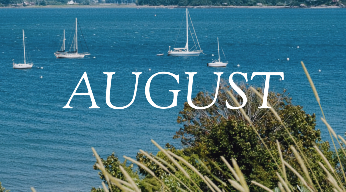 The word "AUGUST" in white text overlaid on sailboats moored in a Maine bay with grasses and trees in the foreground