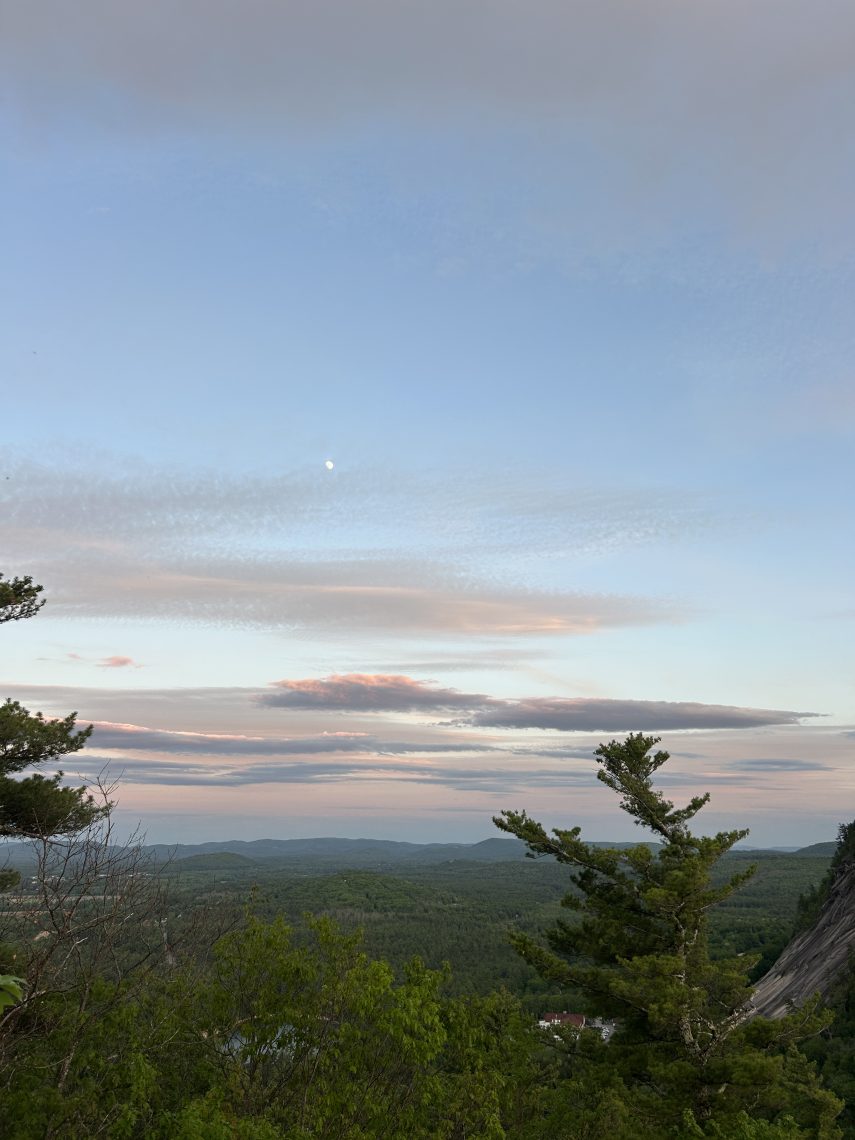 A sweeping sunset view from the top of Cathedral Ledge in Bartlett, NH, looking out over a vast stretch of forested New Hampshire mountains. Pink and purple clouds streak across a soft blue sky, with a faint moon visible above the treeline.