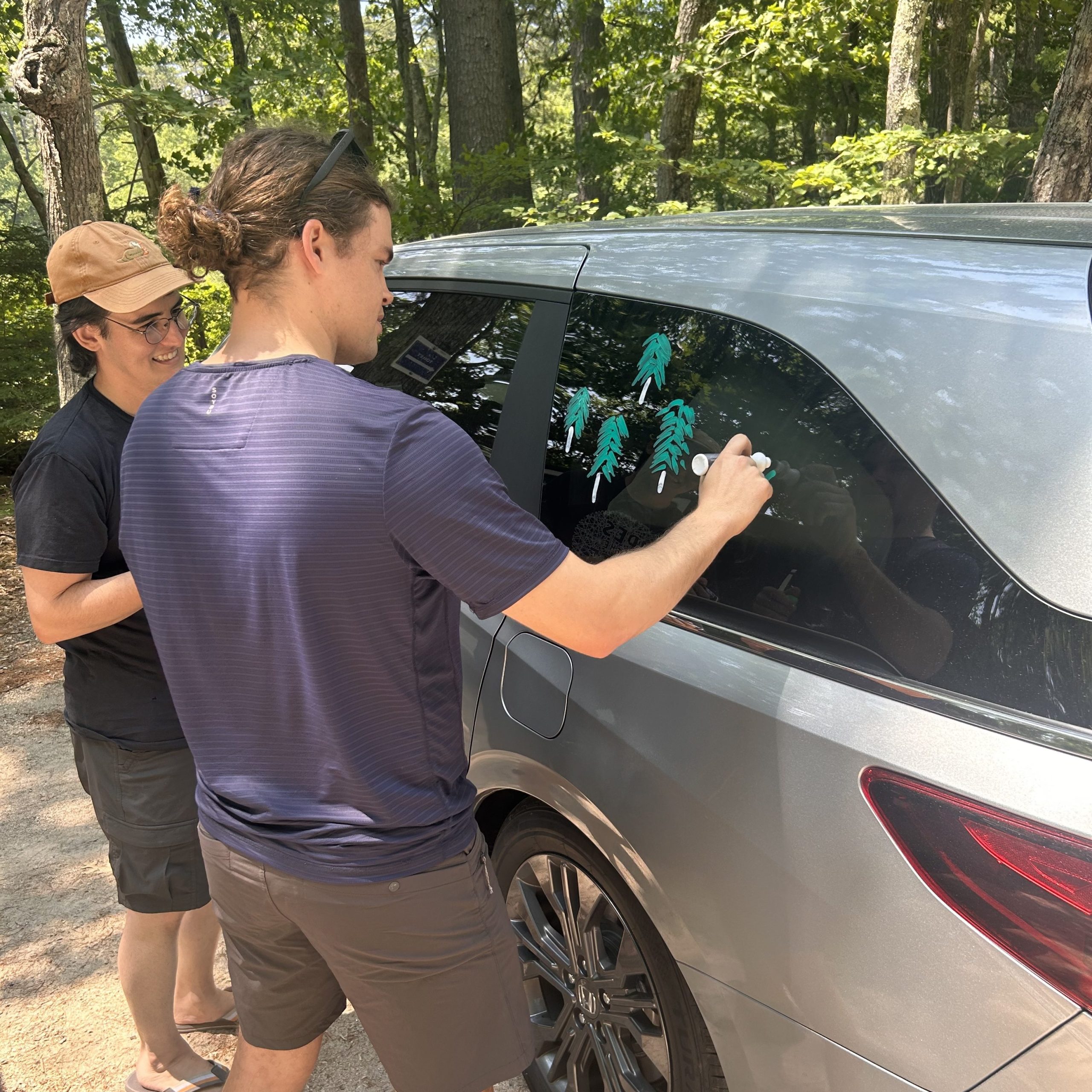 Calvin Soule draws teal pine trees on the Green Curtain Roadshow van window while a fellow participant looks on, parked in a forested area.