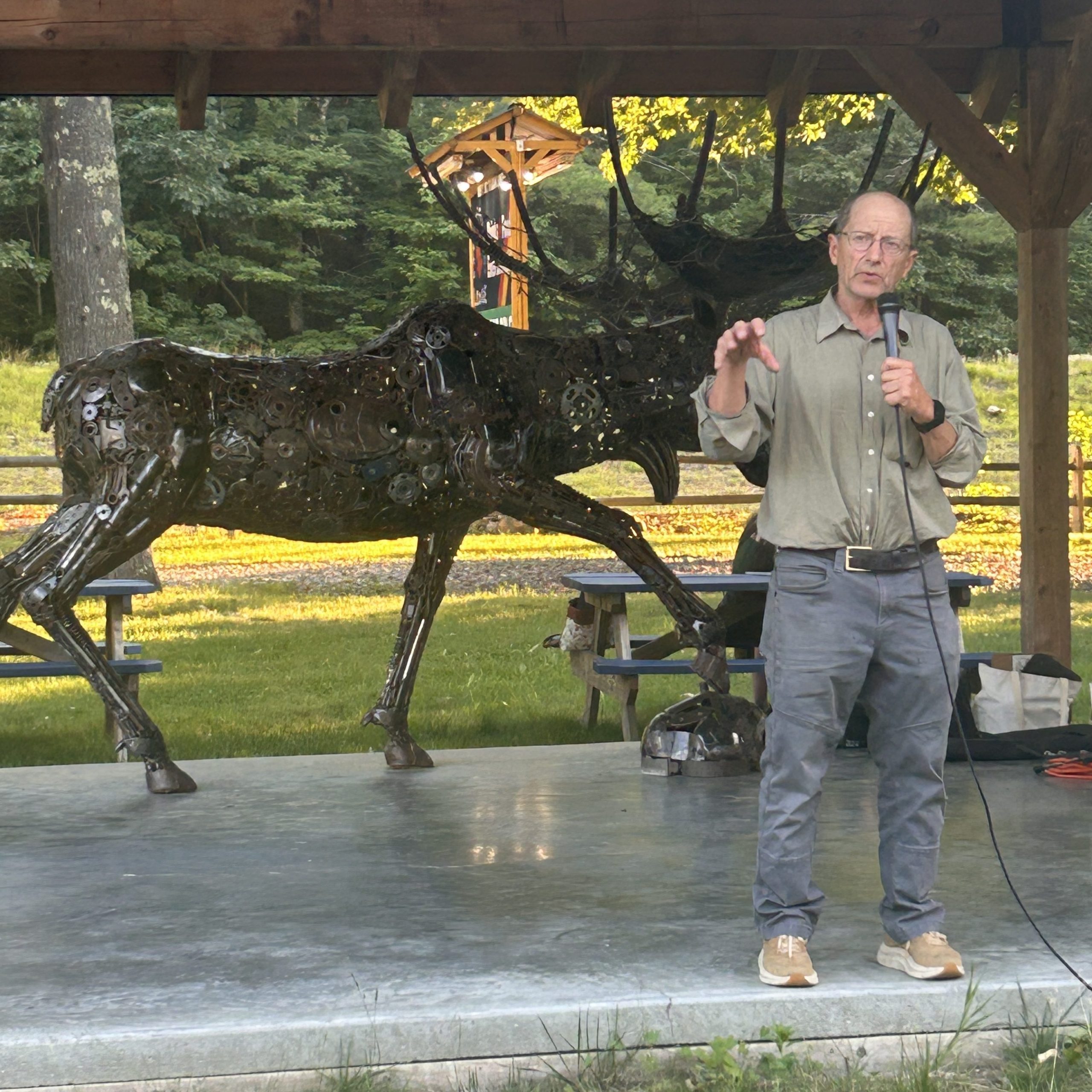 Rick Bass speaks into a microphone at an outdoor event under a pavilion, with a large metal moose sculpture visible behind him.