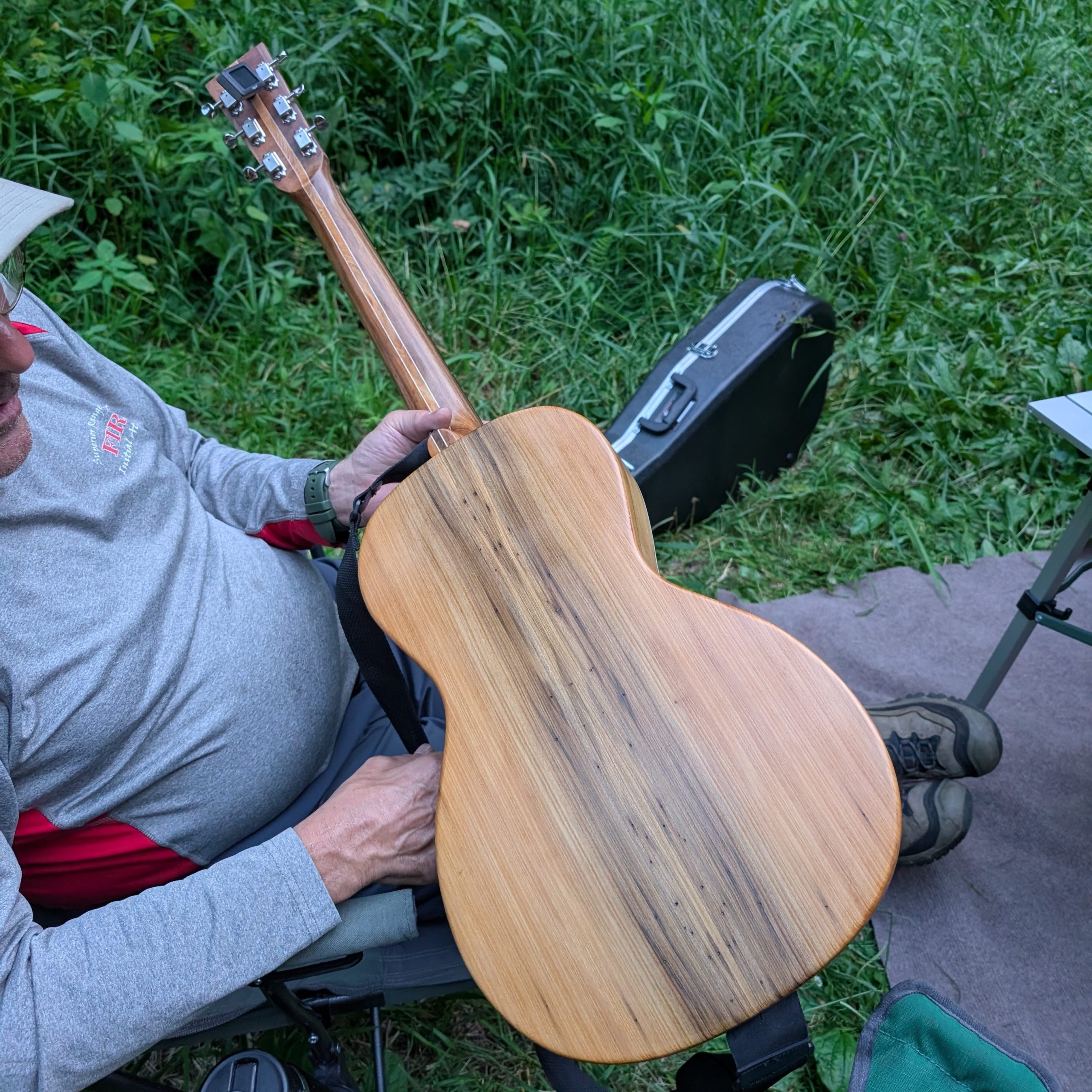 A close-up of the back of the Wormhole guitar, showing its distinctive light-colored wood grain, held by a person sitting outdoors in a camp chair with green vegetation in the background.