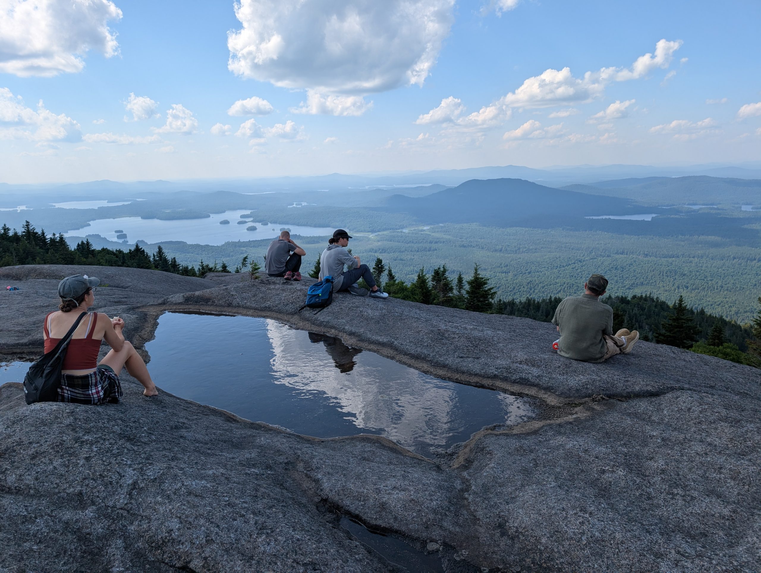 Green Curtain Roadshow participants sit on a granite summit in the Adirondacks, taking in a sweeping view of forested mountains and a lake below, with a small rain pool reflecting the sky in the foreground.