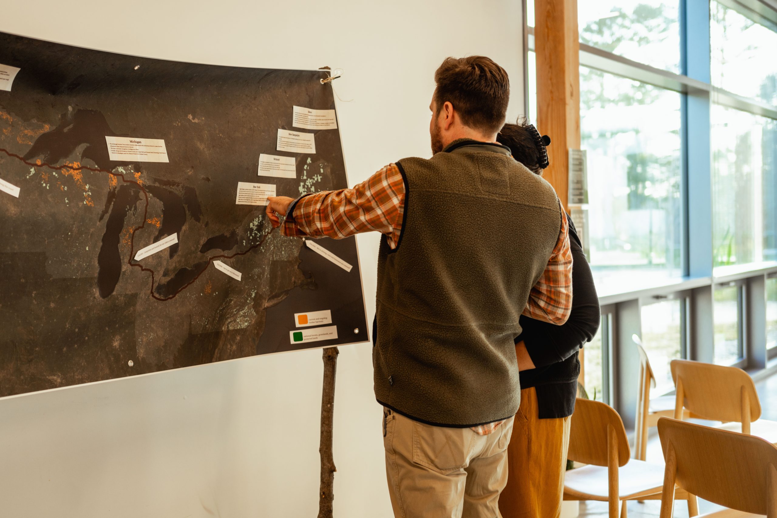 Two event attendees examine the Green Curtain map displayed on a wall, which shows old-growth forests across the northern United States, with orange indicating forests currently being logged and green indicating protected old-growth forests.