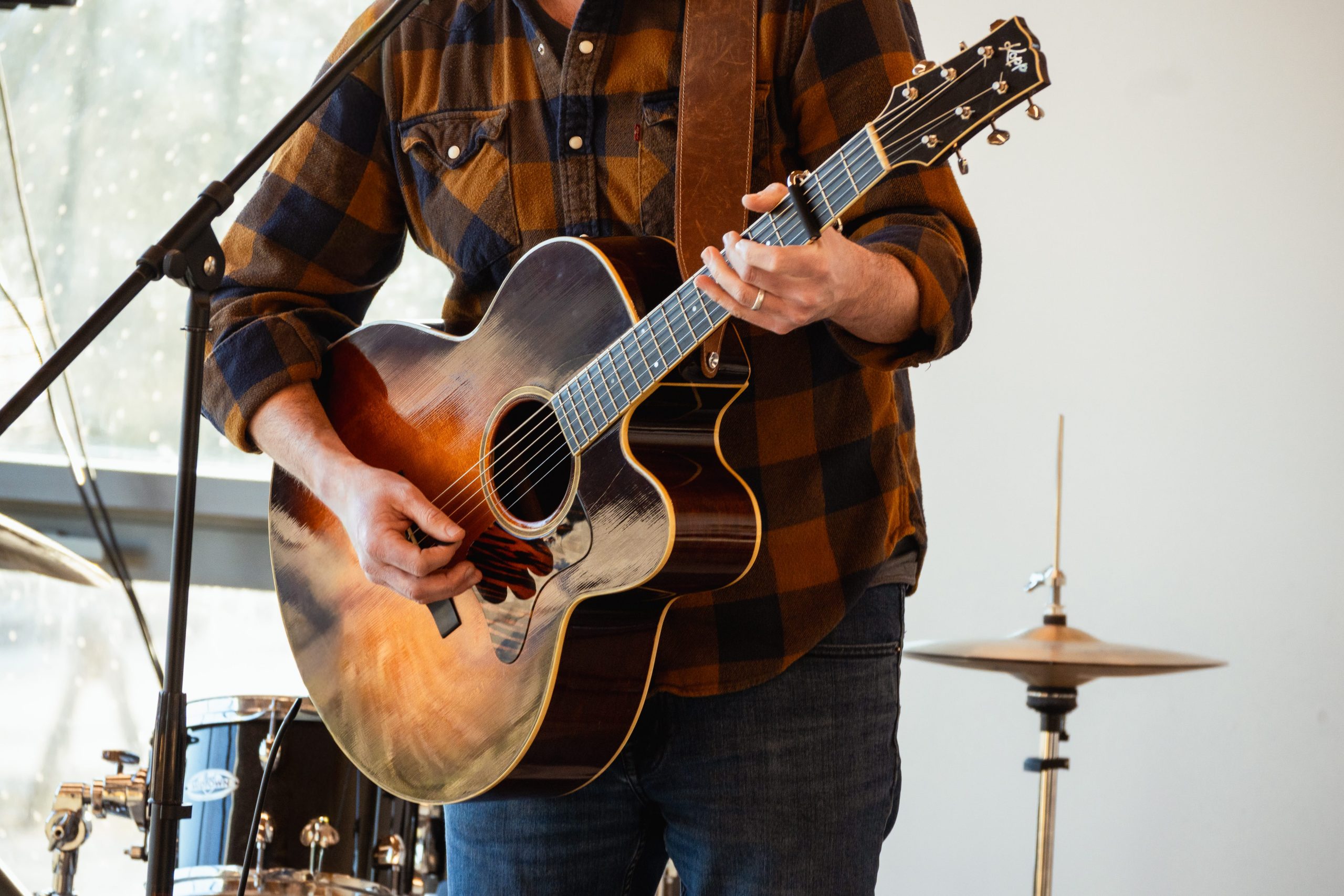 A musician plays the Black Ram Guitar at a live performance, wearing a flannel shirt, with a drum kit visible in the background.