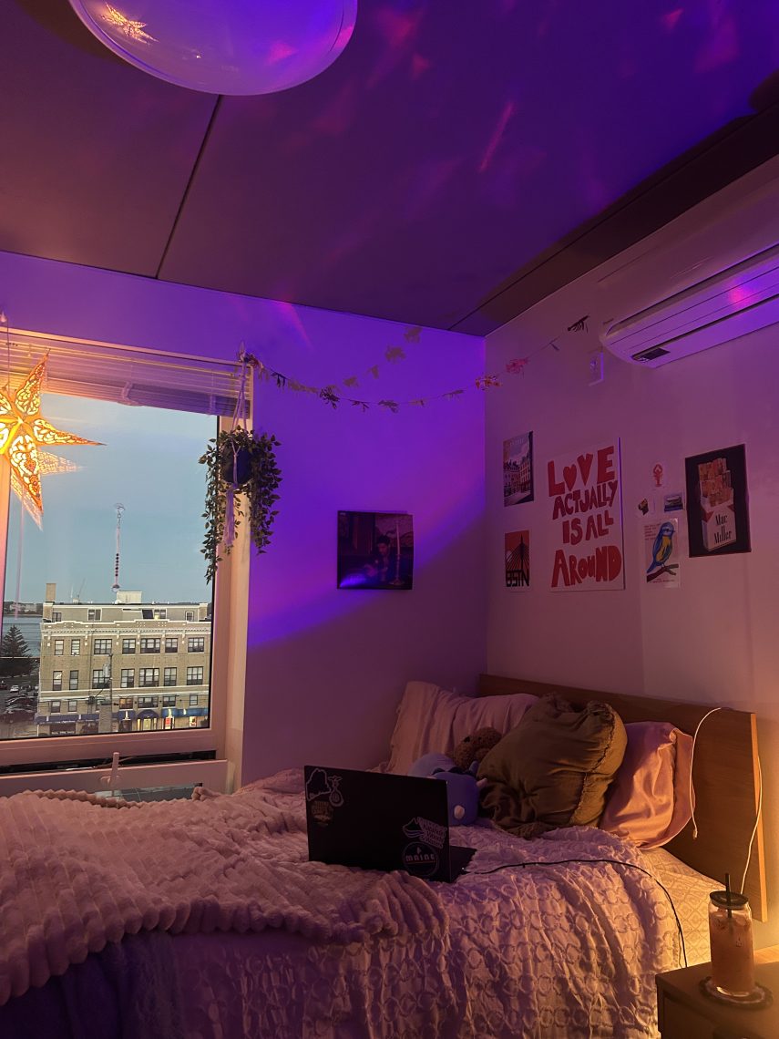 A cozy dorm room glows with purple ambient lighting, with a view of downtown Portland through the window.
