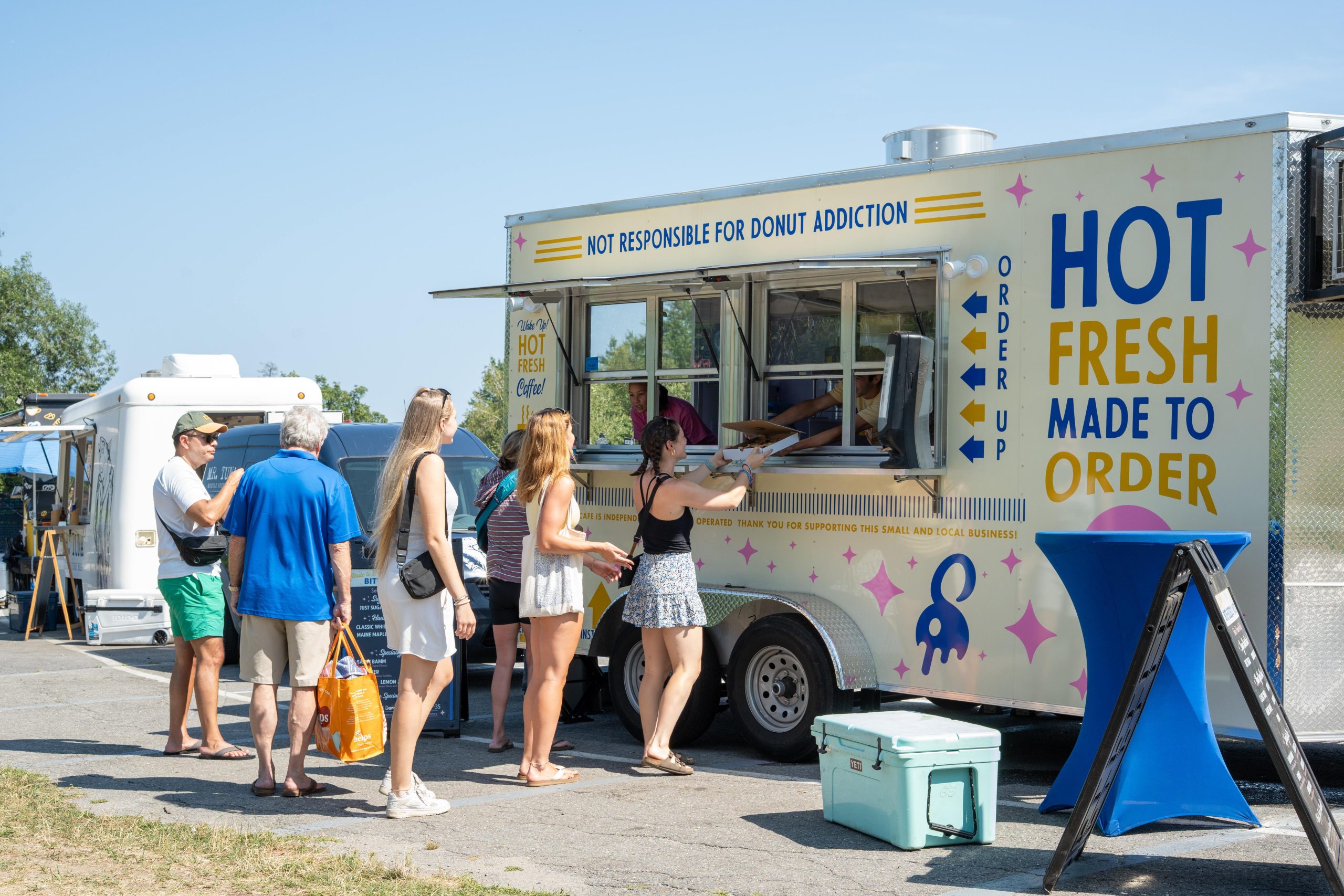 People lined up outside a food truck reading "Hot Fresh Made to Order" at an outdoor summer event