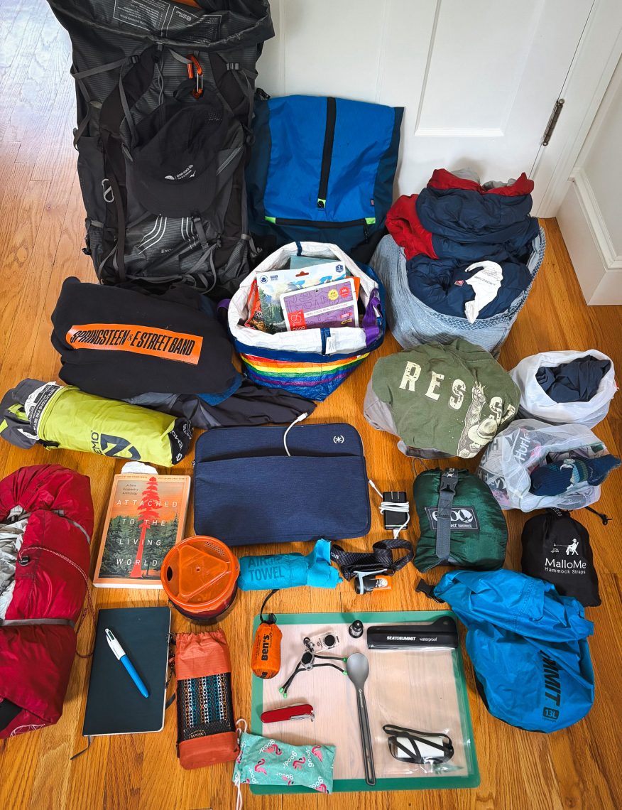 Camping and travel gear laid out on a wooden floor, including a large backpack, sleeping bag, tent, hammock, notebook, laptop sleeve, books, and various outdoor essentials, ready for a two-week road trip.