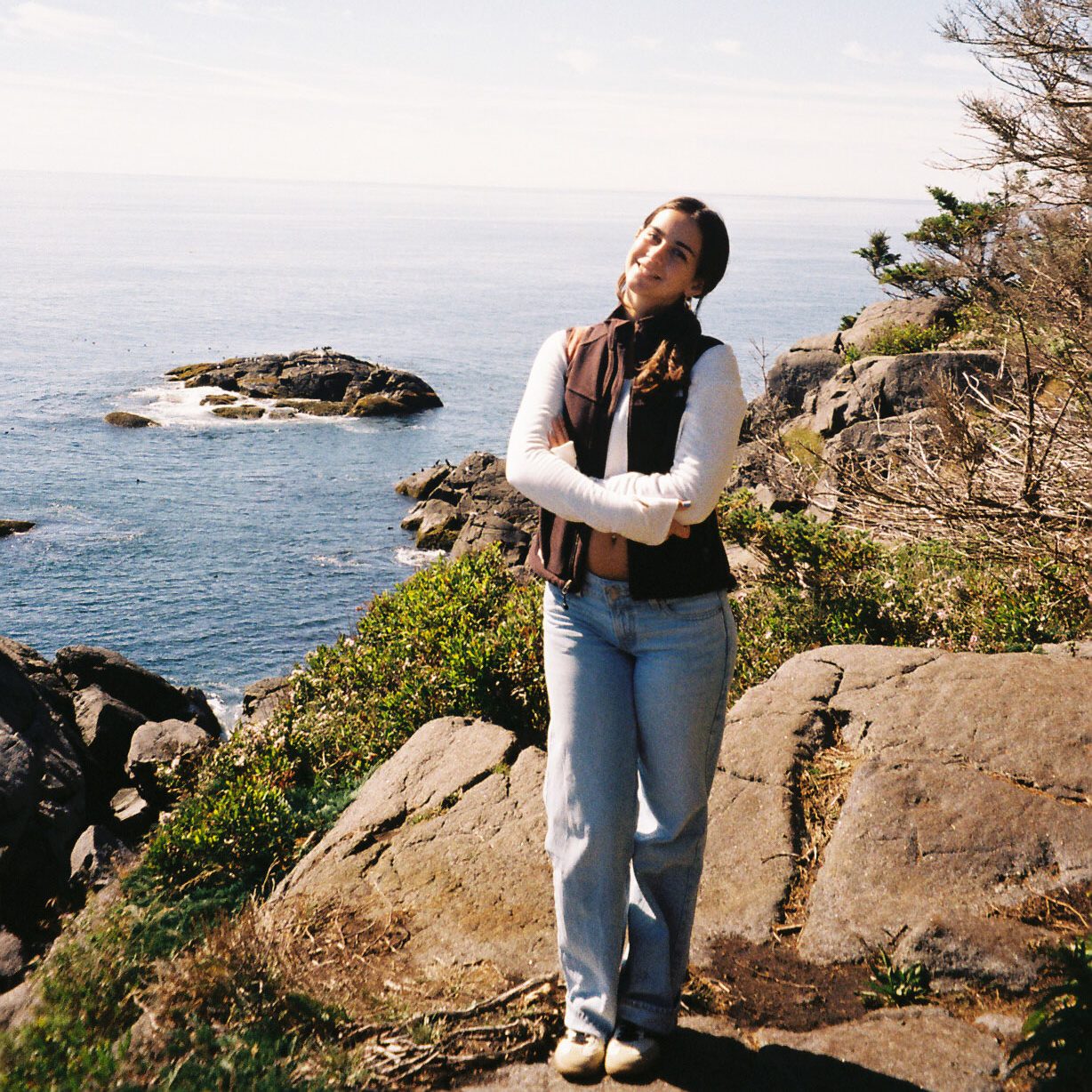 Emma stands on the rocky cliffs of Monhegan Island, ME, arms crossed and gazing upward, with the blue Atlantic Ocean and small rocky outcroppings stretching behind her under a bright partly cloudy sky. Shot on film.