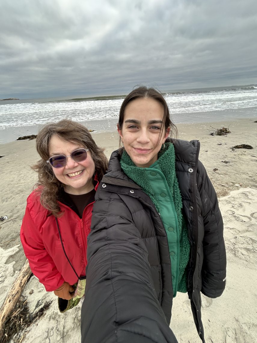 Two people take a selfie on the secluded beach at the end of the Morse Mountain trail in Phippsburg, ME. Both are bundled up in winter jackets, smiling, with a wide sandy beach and dramatic cloudy sky stretching behind them.