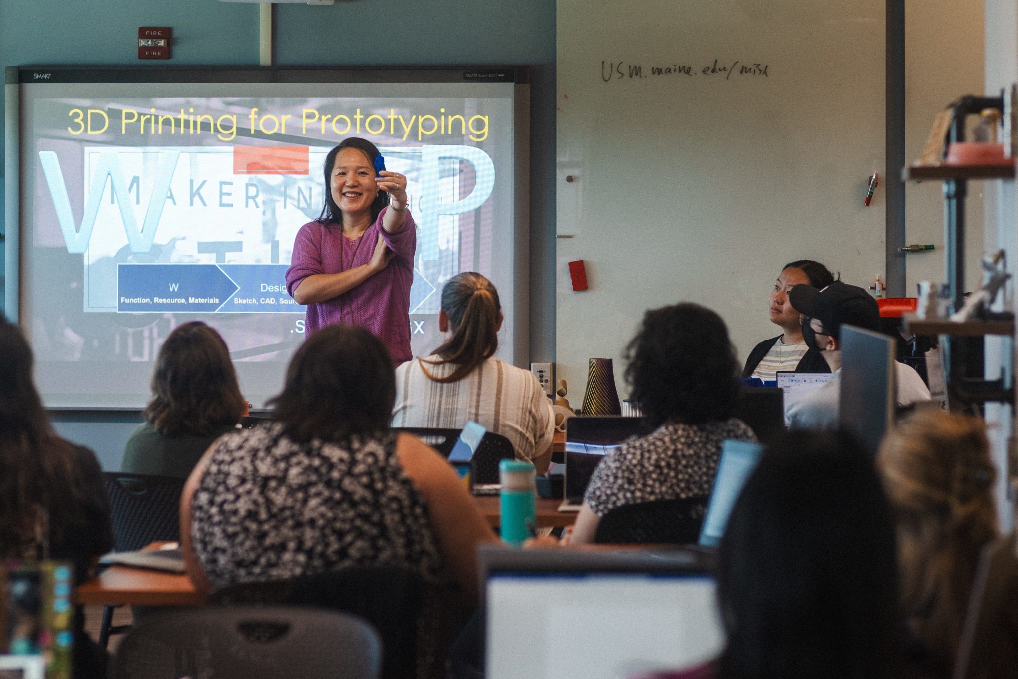 A professor presents a "3D Printing for Prototyping" workshop to a room of seated participants in the MIST Lab, holding up a small 3D printed object while smiling. A smartboard behind her displays a Maker Innovation Studio slide, and the MIST Lab's website address is written on the whiteboard to her right.