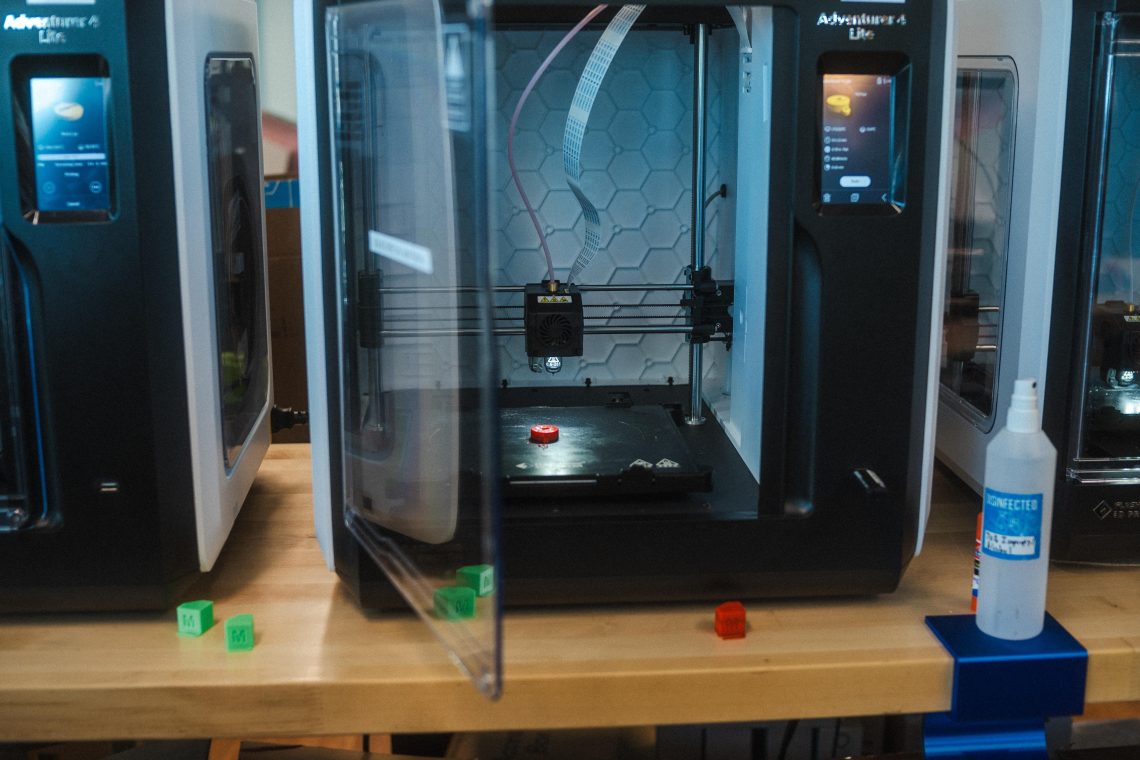 Three 3D printers sit on a wooden workbench in the MIST Lab, one actively printing a small red object. Several small 3D printed cubes are visible on the bench in front of the machines.