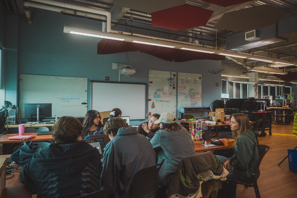 A group of students collaborate around a table in the MIST Lab, with laptops, colorful materials, and project supplies spread across the workspace. Whiteboards covered in notes and equipment stations are visible throughout the open lab space in the background.