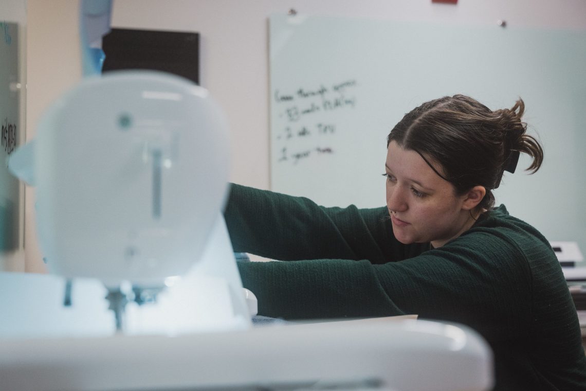 A student leans in to work at a sewing machine in the MIST Lab, focused on her project, with a whiteboard visible in the background.