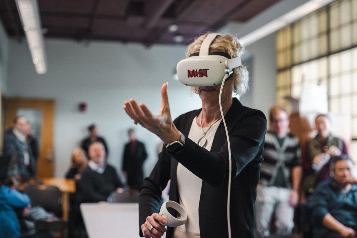 A woman wearing a MIST-branded VR headset reaches out with one hand while holding a controller in the other, surrounded by a crowd of onlookers at a MIST Lab showcase event.