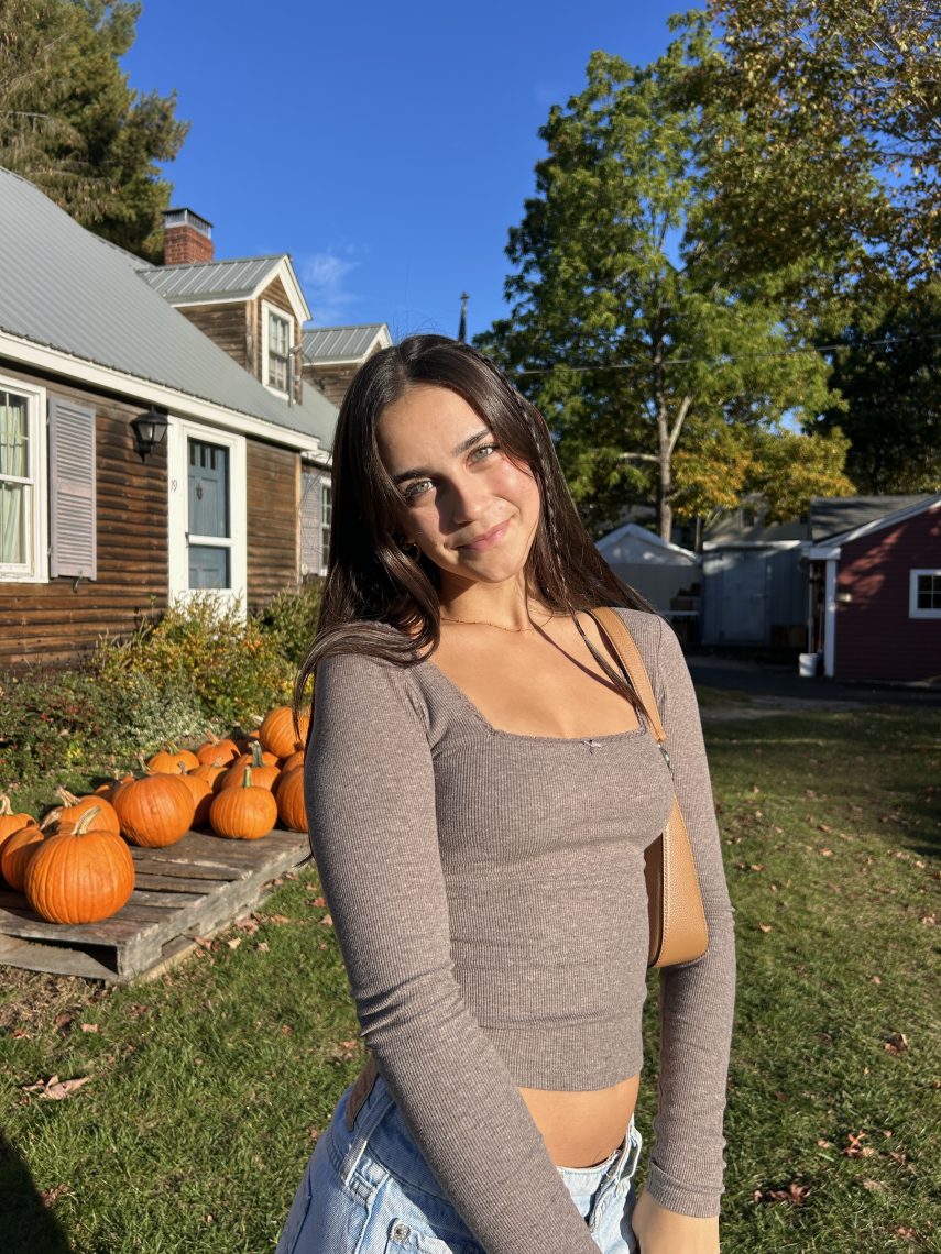 Emma smiles at the camera in front of a display of pumpkins on a sunny fall day.