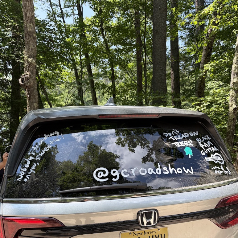 The rear window of the Green Curtain Roadshow's Honda van, decorated with hand-drawn messages and drawings in white and teal marker, including '@gcroadshow', 'Don't Tread on Trees', 'Black Ram Guitar', 'Nobody's Standing on You, Sweet Pie', and a small illustrated tree, parked in a forest setting with a New Jersey license plate visible.