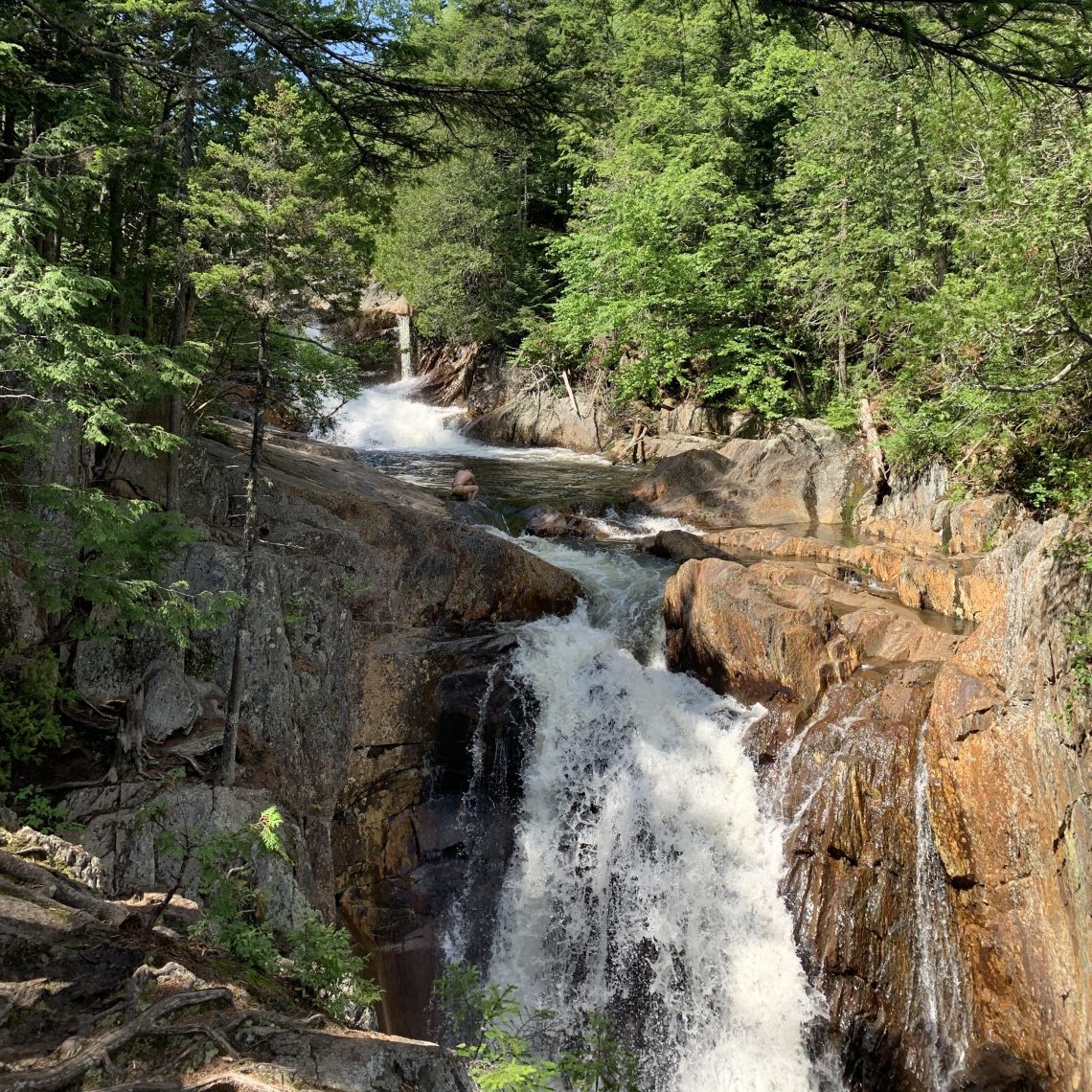 Smalls Falls in Rangeley, ME rushes down a series of rocky ledges surrounded by dense green pine trees under a clear blue sky. Natural pools are visible at the base of the falls.