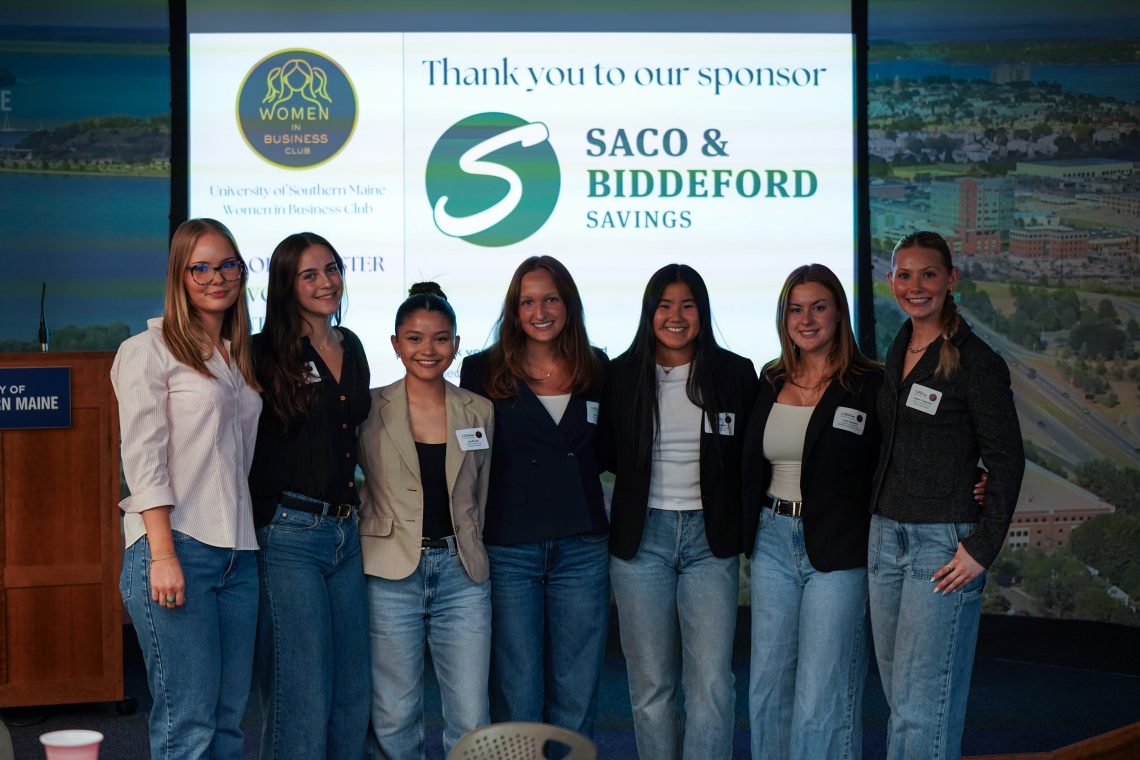 Seven members of USM's Women in Business Club pose on stage in front of a screen thanking sponsor Saco & Biddeford Savings.