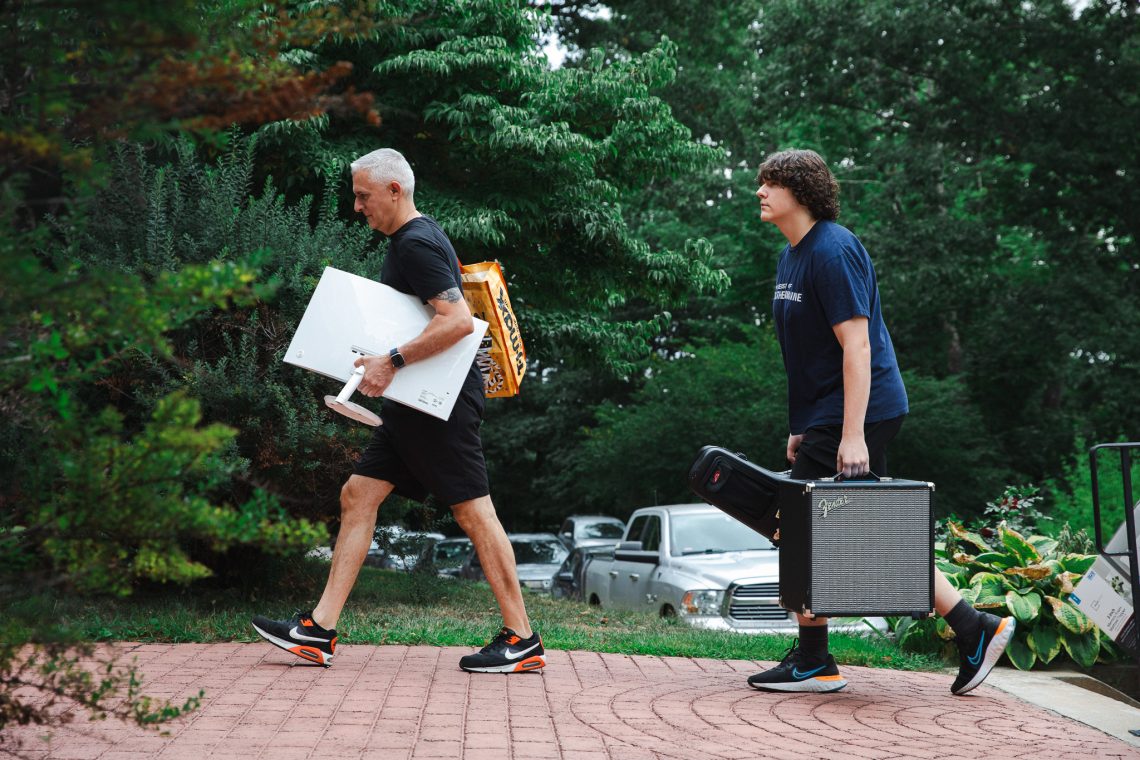 Father and son carrying a monitor and guitar amp while walking on a brick path during move-in day