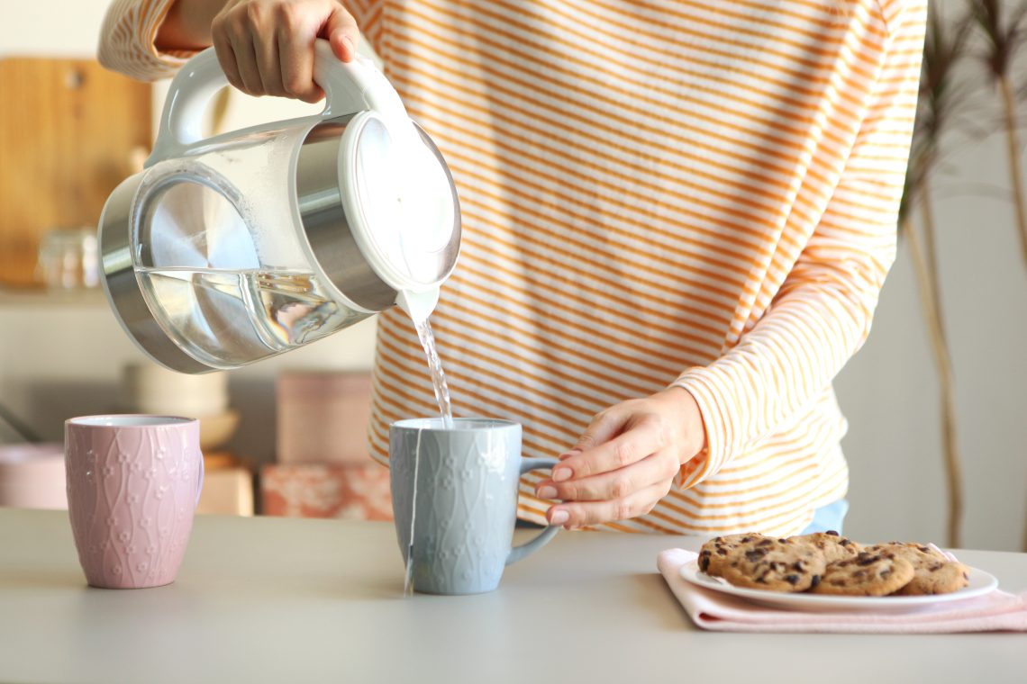 Person pours hot water from an electric kettle into a mug next to a plate of cookies