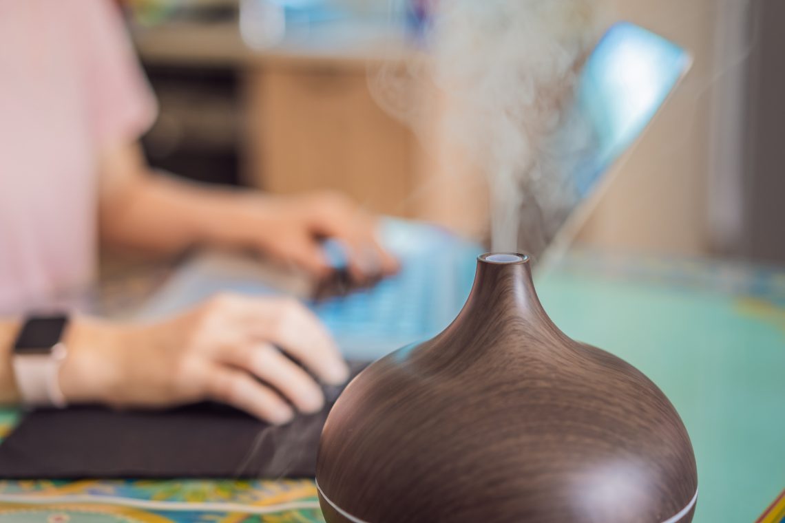 Wood-grain essential oil diffuser releasing mist on a desk while someone works on a laptop in the background
