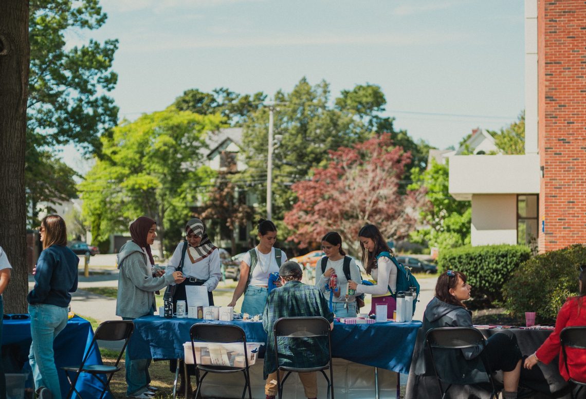 Students gather around outdoor tables set up with informational materials and giveaway items on a sunny campus day.