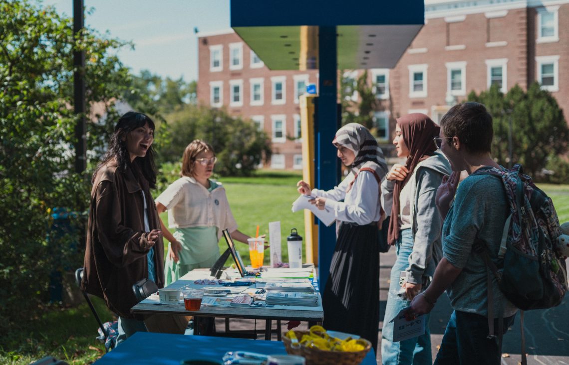 Students stop to chat with representatives at a club or resource table during an outdoor fair on USM's Portland campus. A group of students reviews materials while a staff member engages with them enthusiastically, with the large blue USM "T" sculpture visible in the background.